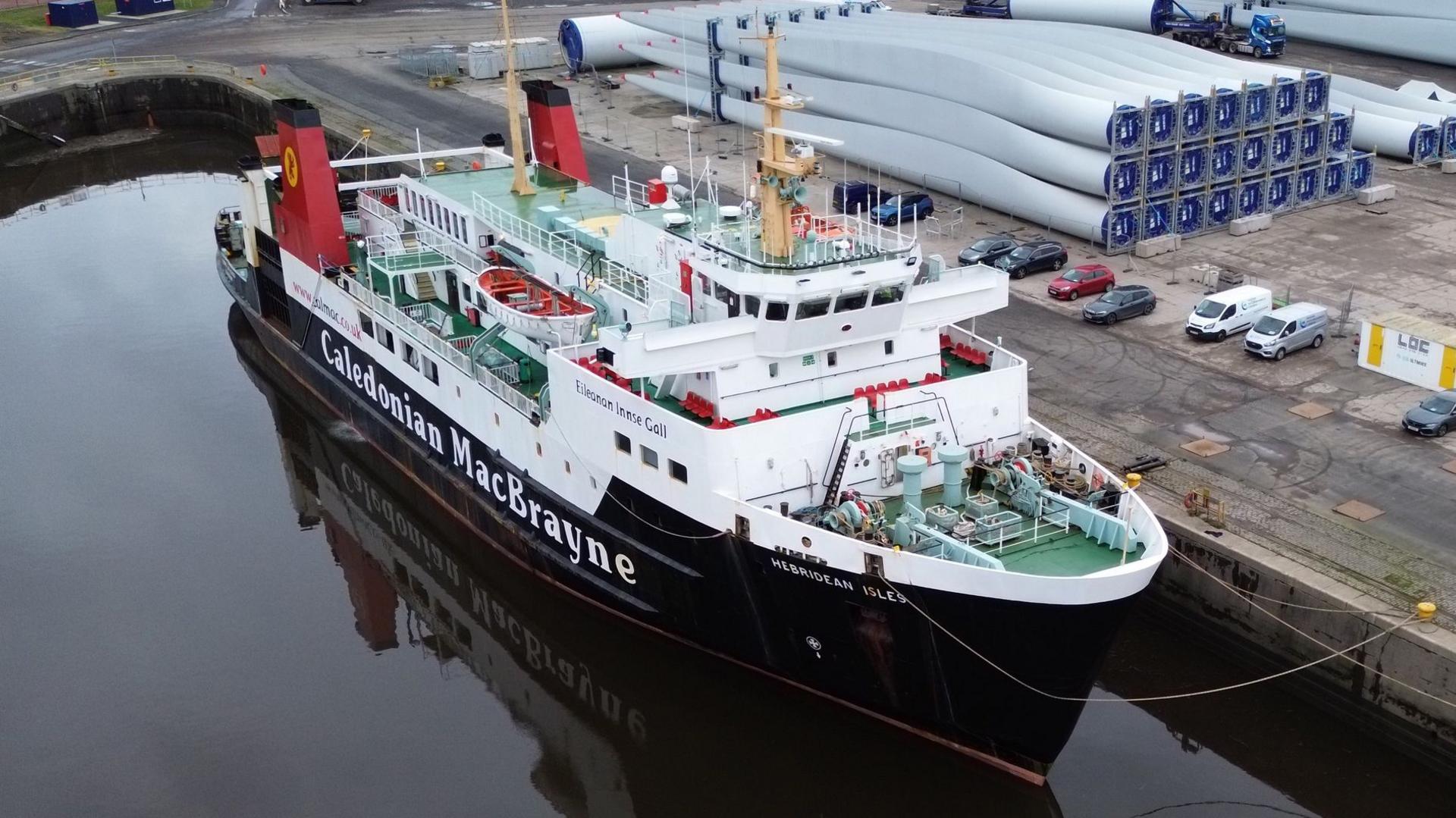 A large black and white ferry with red funnels, moored at a quayside with wind turbine blades seen on the wharf beside it