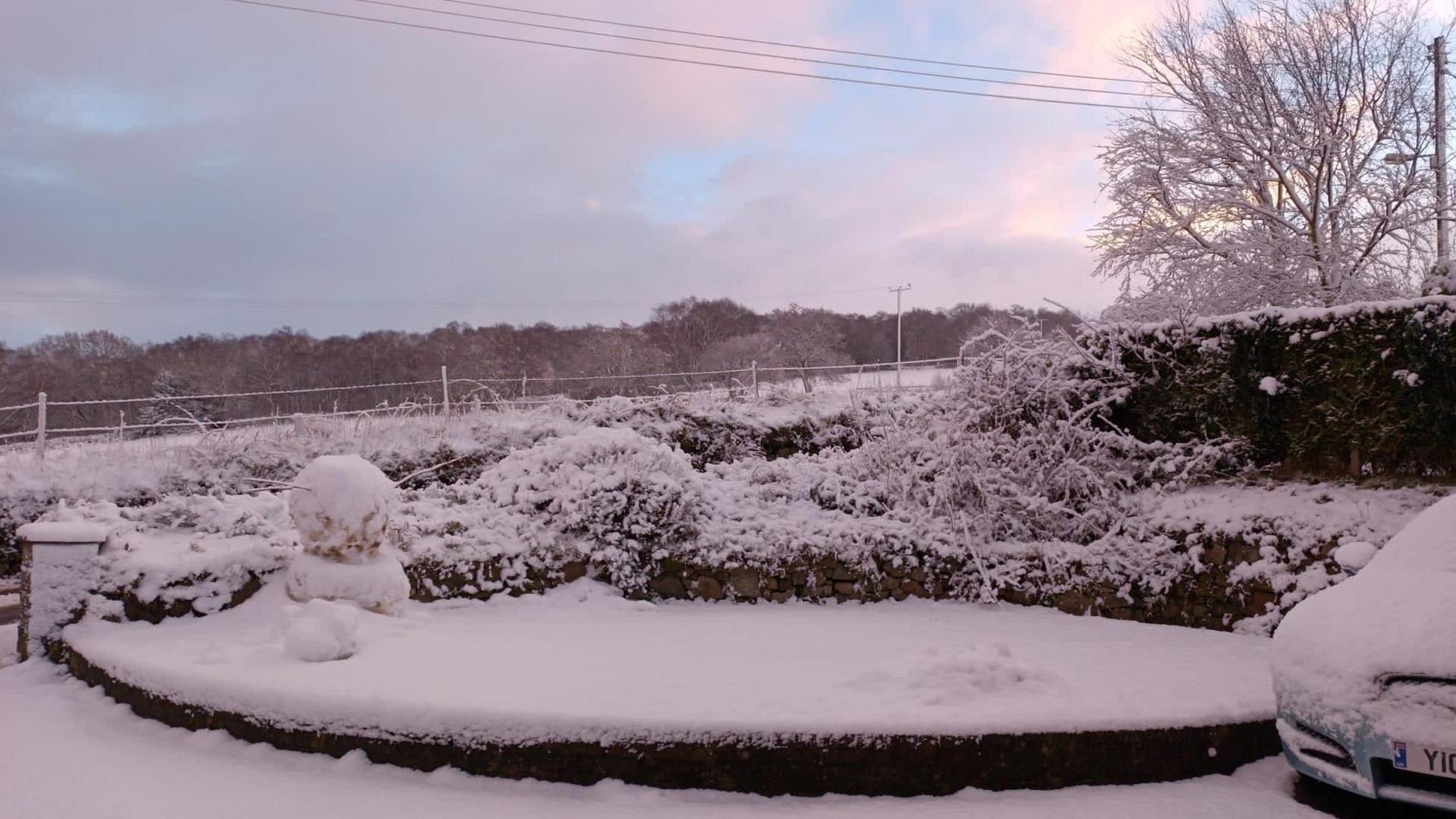 Snow in Mow Cop. A garden with a snowman and snow on hedges and on top of a car. The sky is cloudy with some blue sky also in view. 