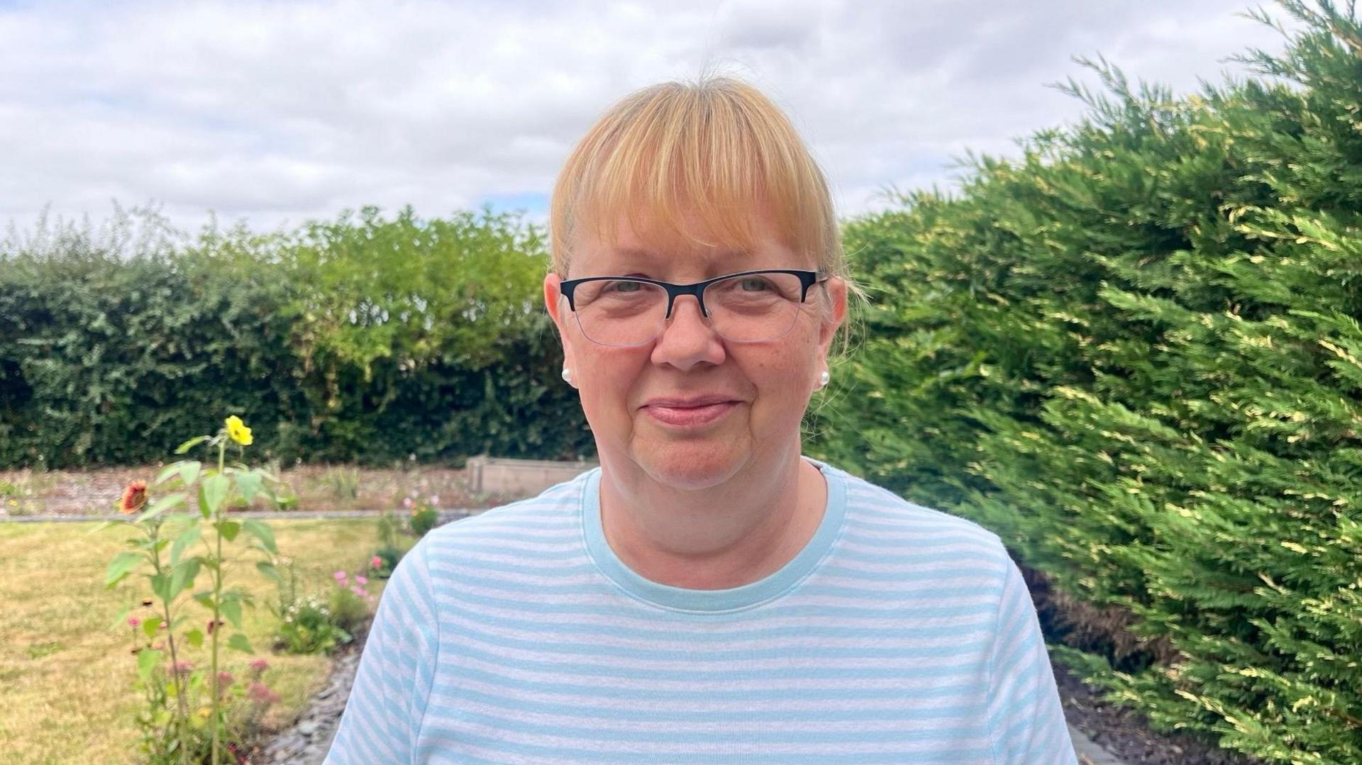 A woman - Janet Blessington - stood in a garden with bushes surrounding her. She has a ginger fringe and is wearing glasses, pear earrings, and a blue and grey striped t-shirt.