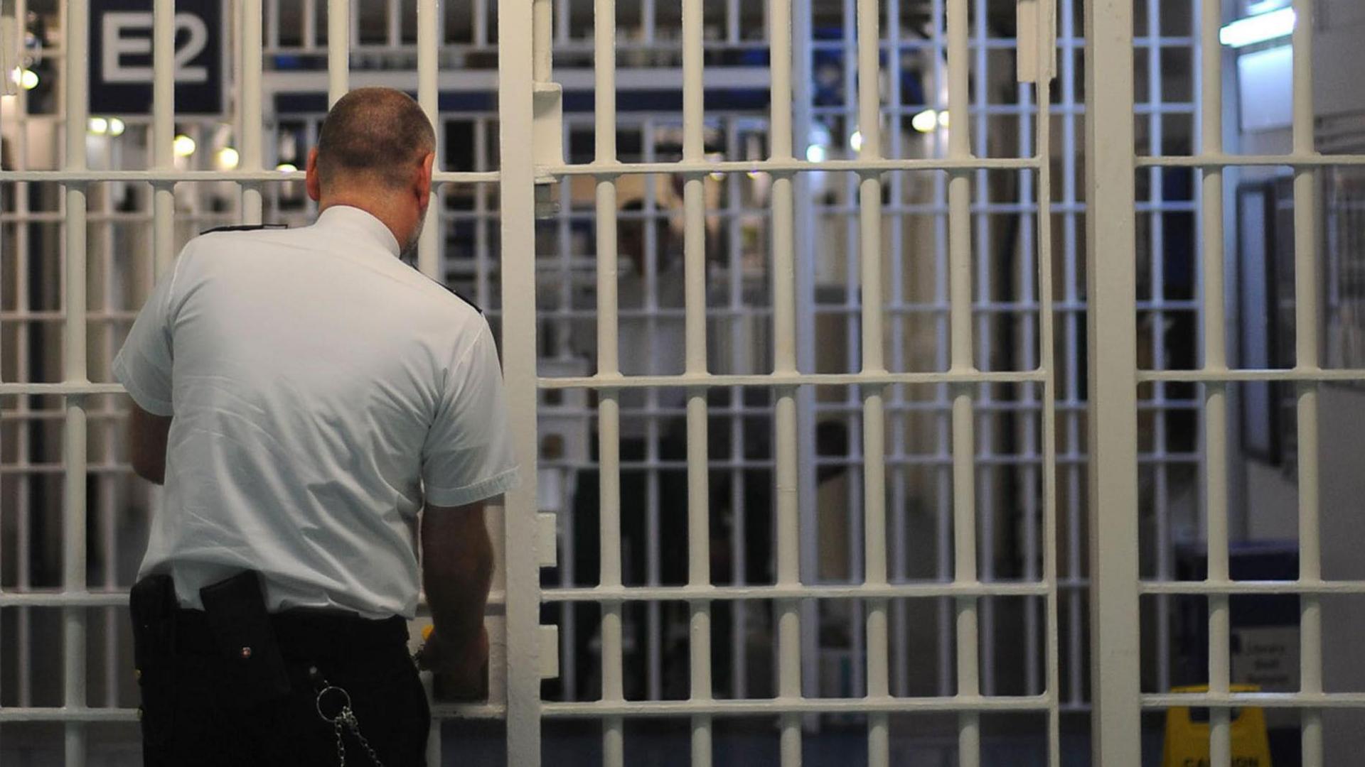 A prison officer, wearing a white shirt and dark trousers, stands with his back to the camera at a barred gate. The vertical and horizontal bars are white and another gate can be seen behind them.