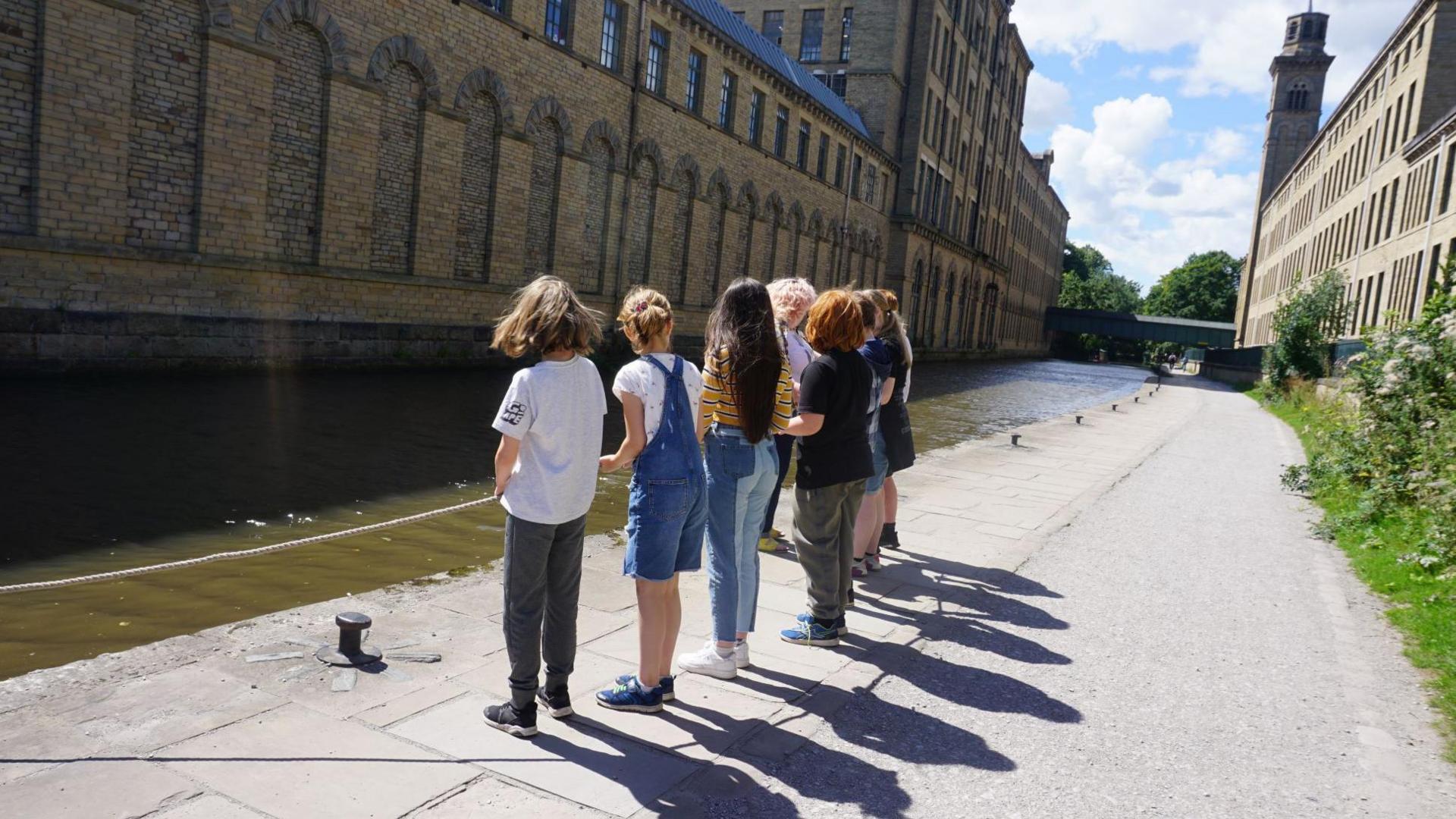 A group of young people stood at the side on a river on a sunny day. On one side of the river is a large building. 