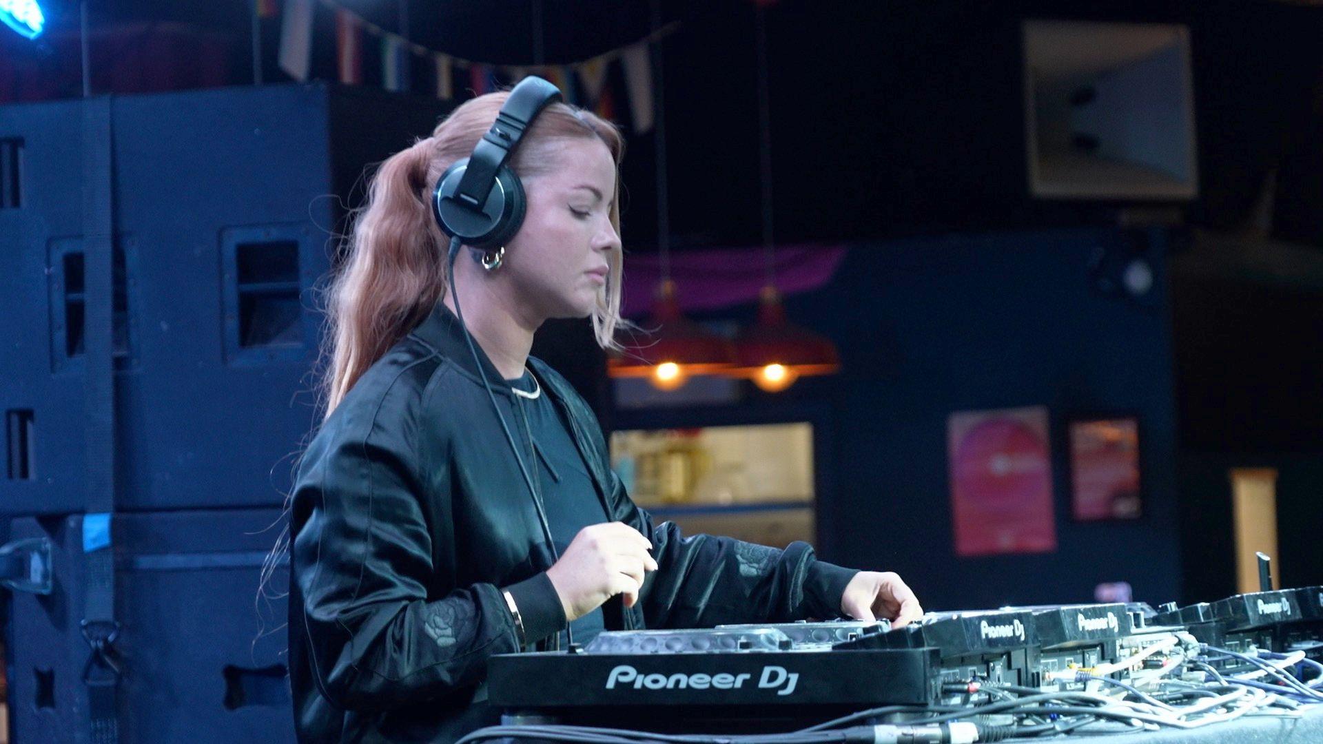 Harriet Jaxxon is sound mixing with DJ decks. She is wearing black wired headphones and a black bomber jacket and T-shirt