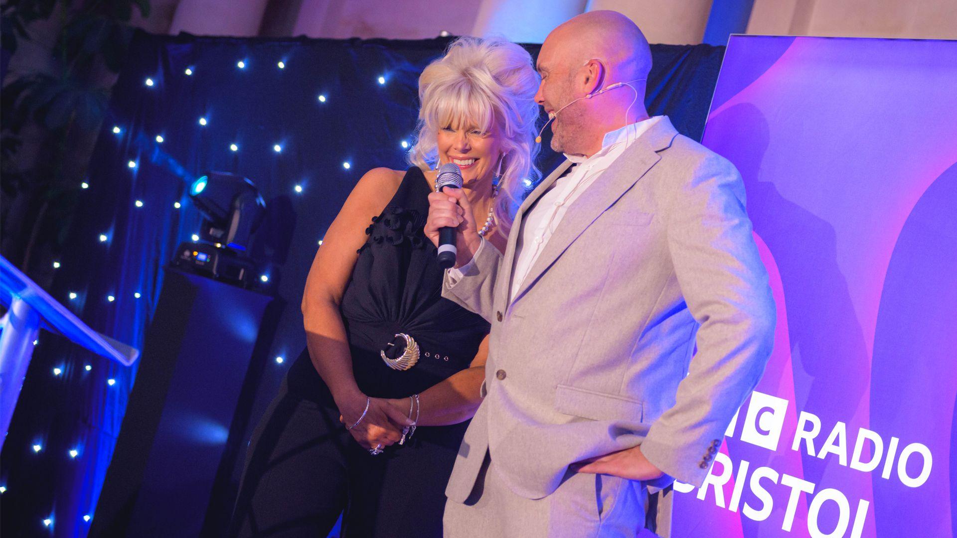 A woman with bleach blonde hair tied up and wearing a black jumpsuit smiles at the audience. Presenter Joe Sims, wearing a grey suit, is holding a microphone to her mouth. Purple BBC Radio Bristol branding is behind them onstage.