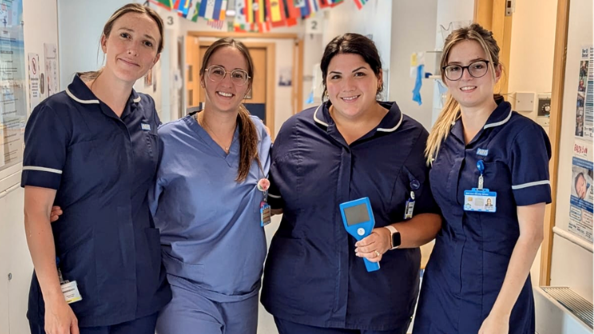 Four female nurses stand together in a hospital corridor. They are wearing blue scrubs.