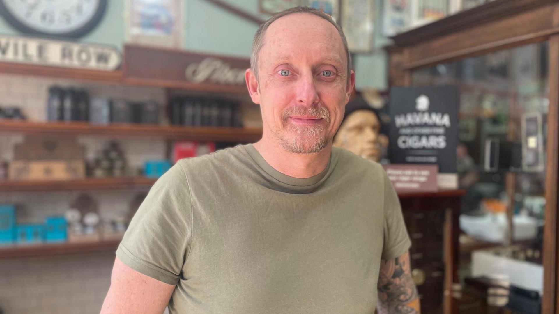 A man with cropped hair, a beard and a green t-shirt stands in front of a counter at a barber shop