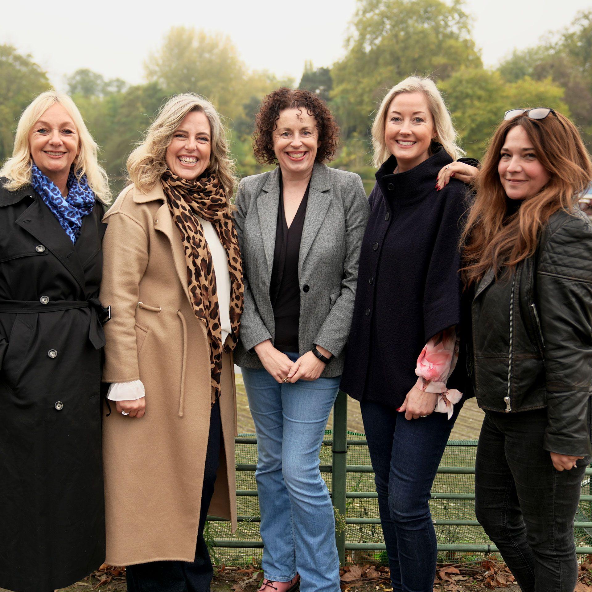 Five women, wearing winter jackets and scarves, standing together in a park on an autumn day. All five are smiling at the camera.