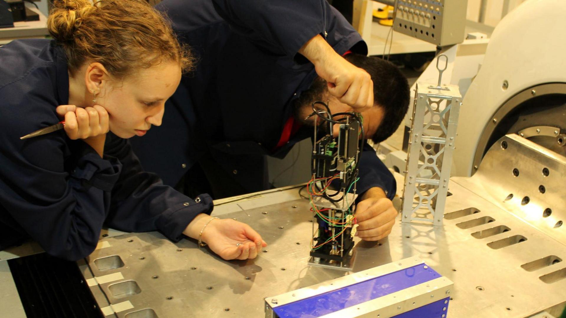 Two students working on a satellite which is covered with wires