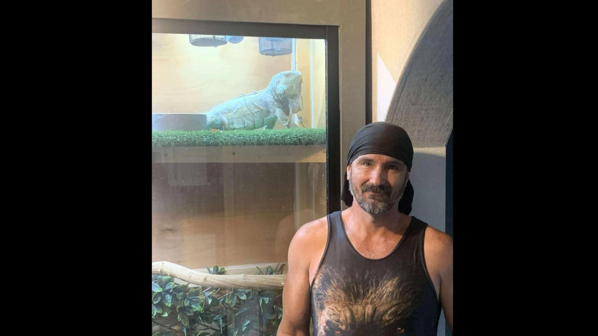 A man in a dark grey singlet and dark grey bandana is standing in front of a vivarium. A blue-coloured iguana is inside the enclosure.