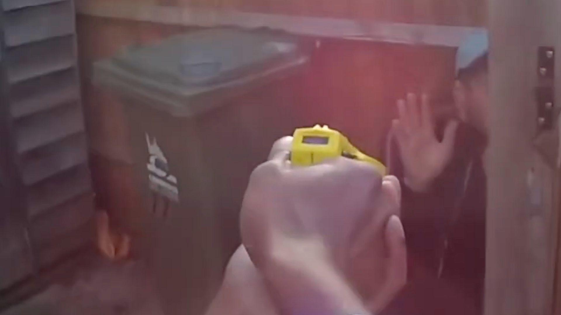 Hands holding a taser gun and pointing to a man hiding among some wheelie bins