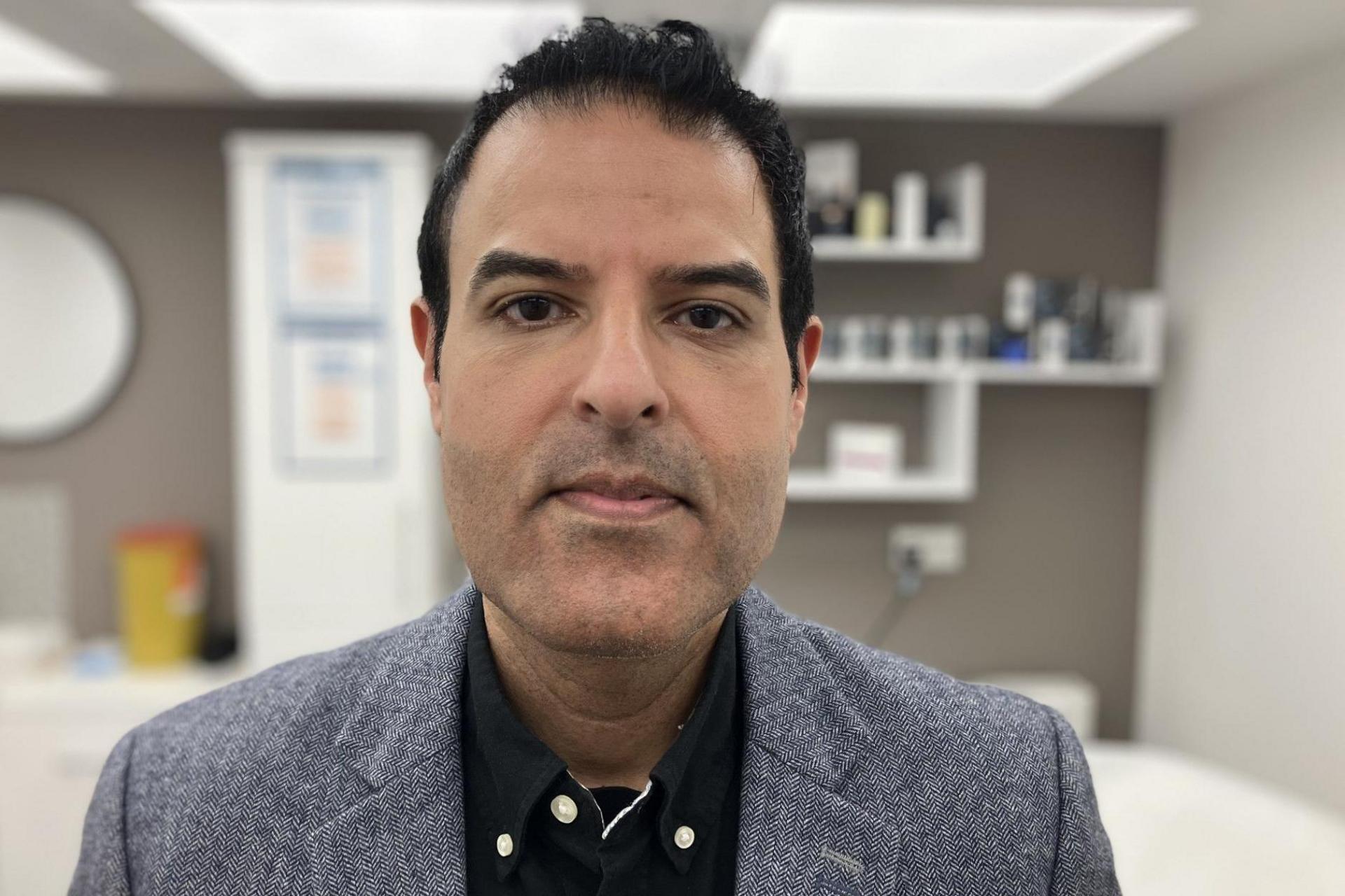 The image is a close-up portrait of a person positioned centrally in an indoor setting. The person is wearing a textured grey blazer over a dark button-up shirt and is facing directly toward the camera. The background is softly blurred and appears to be a clean, modern office, clinic, or consultation room with shelves, framed items, and various small objects visible. 
