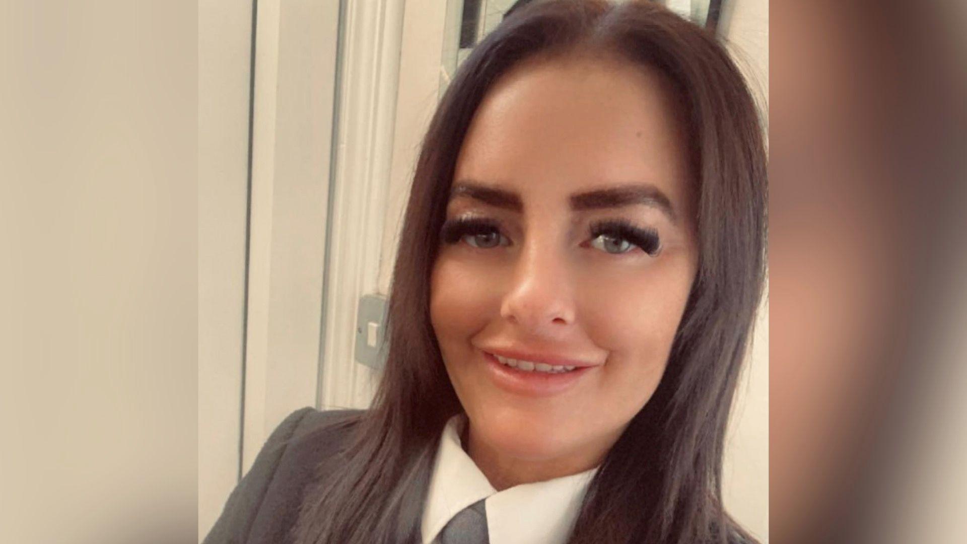 A woman with straight dark hair smiles at the camera. She is wearing a grey blazer and white collared shirt.