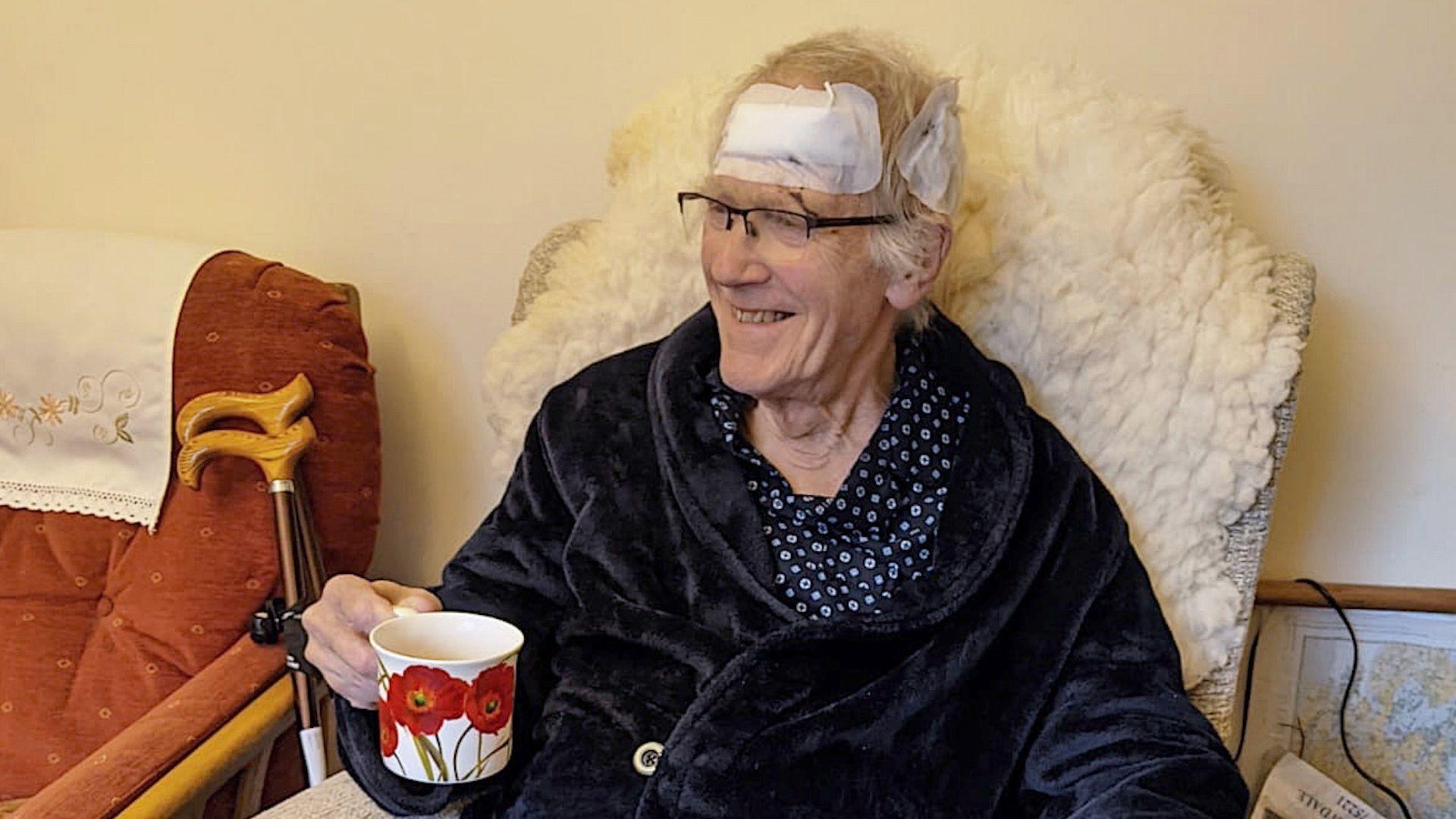 Brian Mico sitting in a chair wearing his pyjamas and dressing gown, holding a cup of tea. Mr Mico is smiling but has two large white padded dressings on his head after injuries sustained during a fall on the pavements.