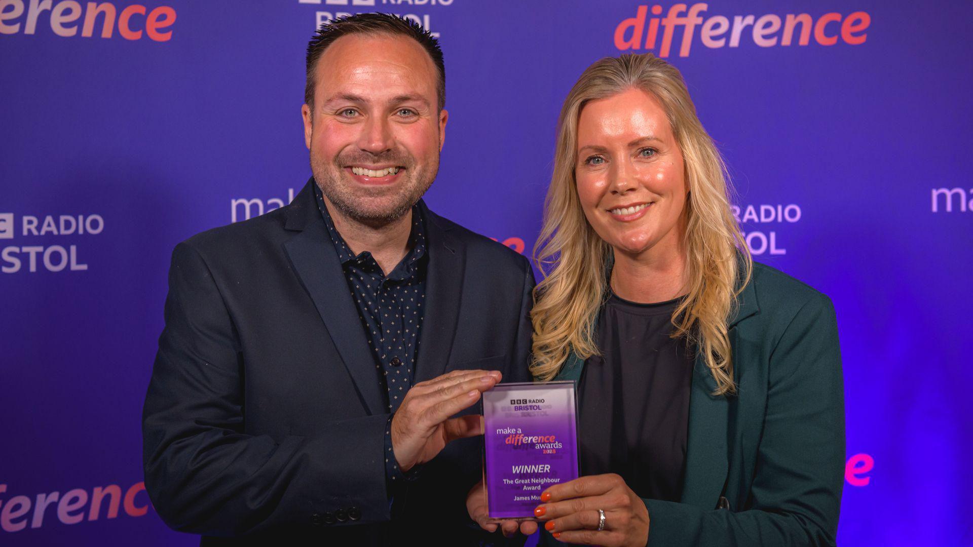 A man wearing black suit smiles at the camera next to blonde woman in a green blazer whilst holding a winner trophy in the centre. The background is purple.