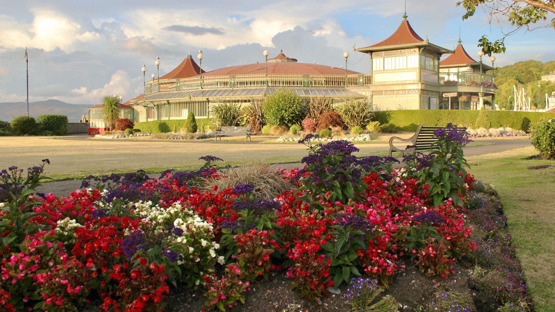 A park with a Victorian-era pavilion in the background. Colorful flowerbeds are in the foreground, and the sky is filled with dramatic clouds.