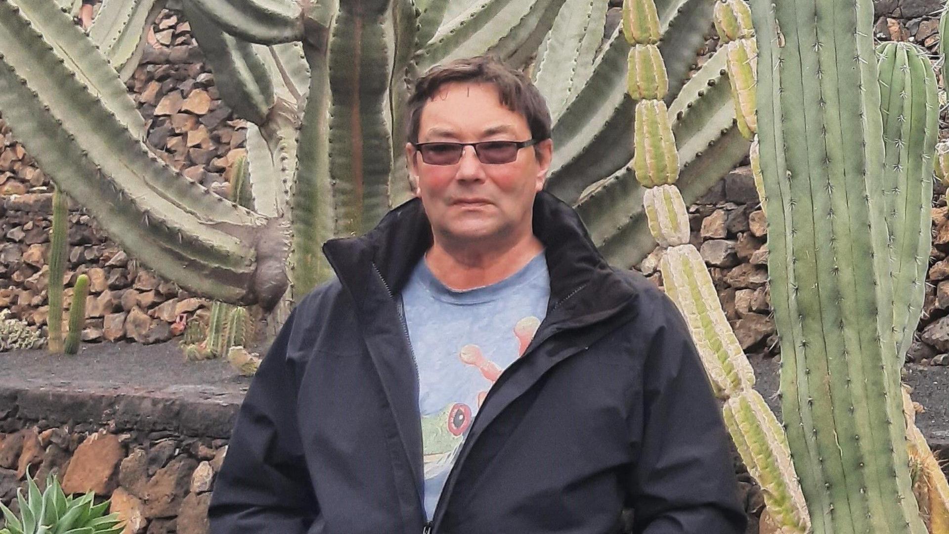 Frank Ingram, pictured wearing a black rain coat and a light blue/grey t-shirt with a green frog on the front. He has dark hair and is wearing dark glasses. He is unsmiling, and seen standing in front of a brick wall and a number of very large cacti. 