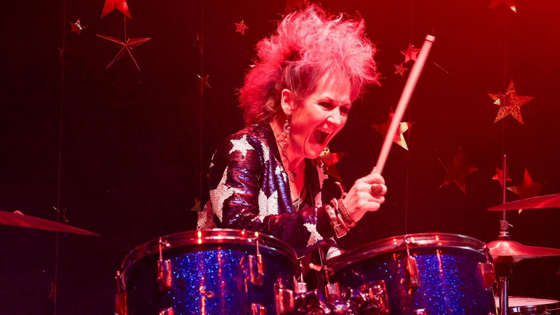 Lorraine Ashbourne on stage playing the drums. She is wearing a dark blue sequin jacket patterned with white sequin stars. She is holding a drum stick in her right arm as she prepares to hit the sparkly, blue kit. She has bright pink hair and has her mouth open wide.