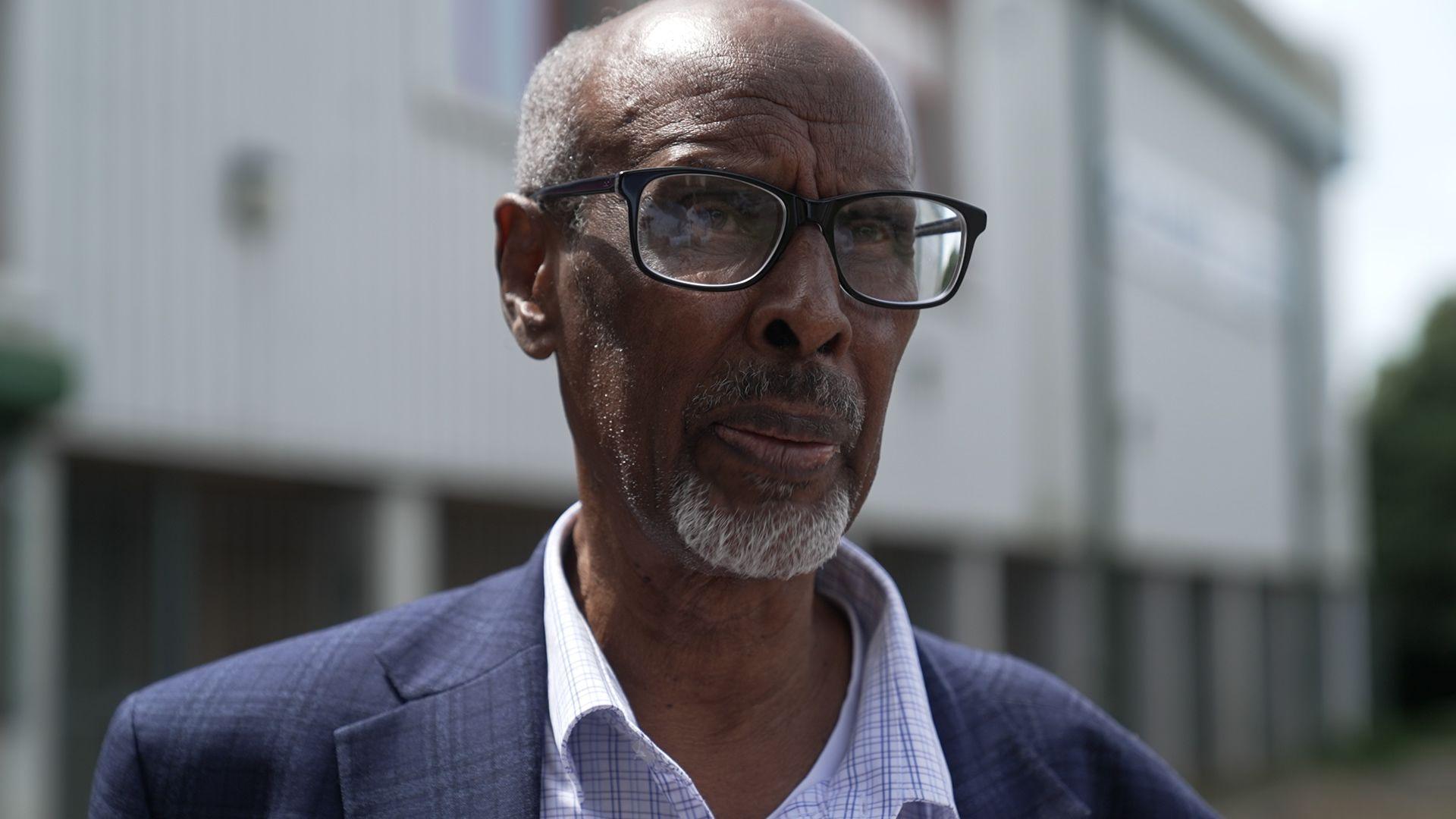 Eid Ali Ahmed stands outside the conference venue. He's smartly dressed in a navy suit, and black rimmed glasses. He has a neatly trimmed grey beard and cropped white hair.