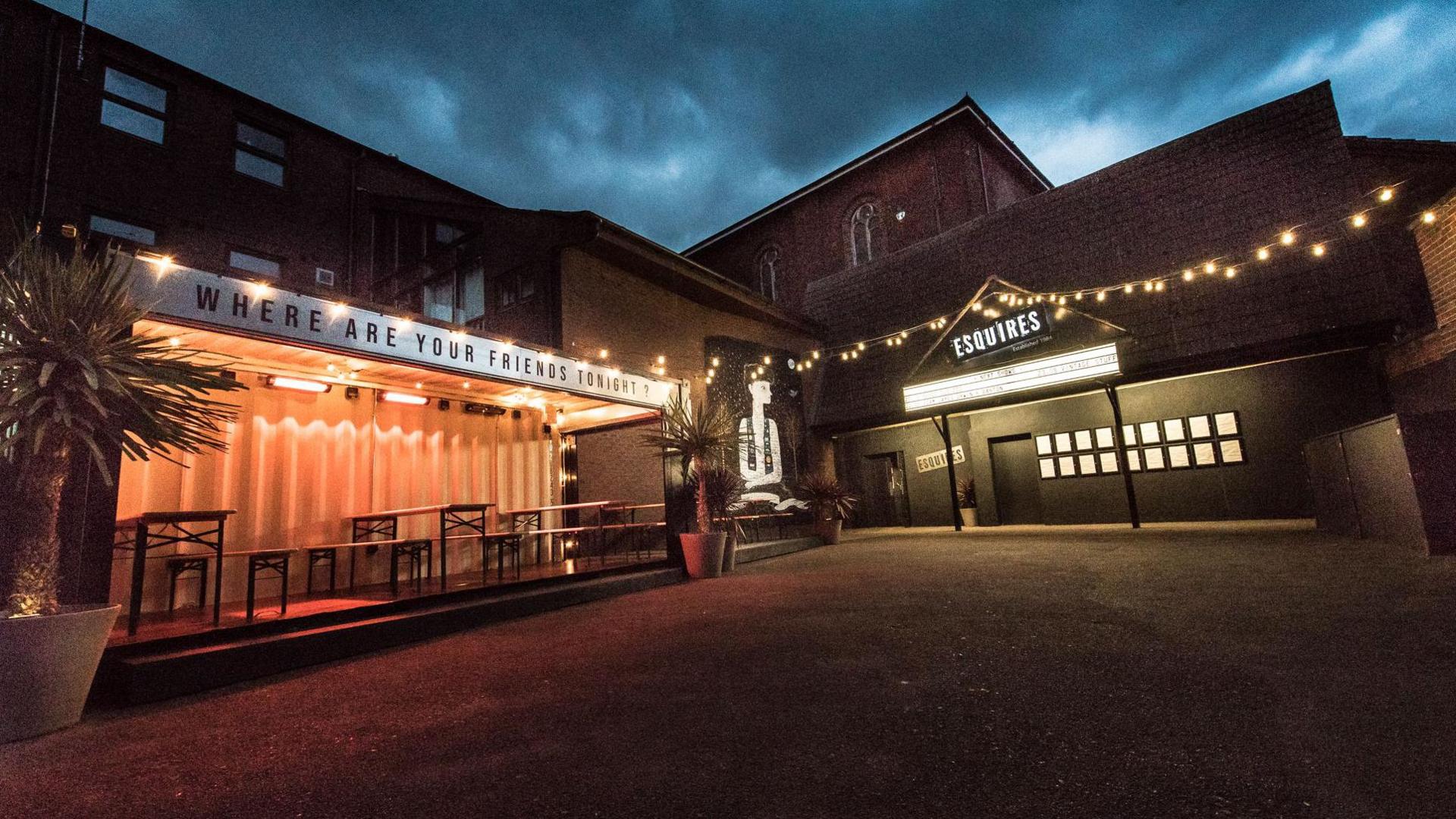 A view of Bedford Esquires at night. It has outdoor lightbulbs lining the courtyard. There is a seating area outside made out of a former shipping container.