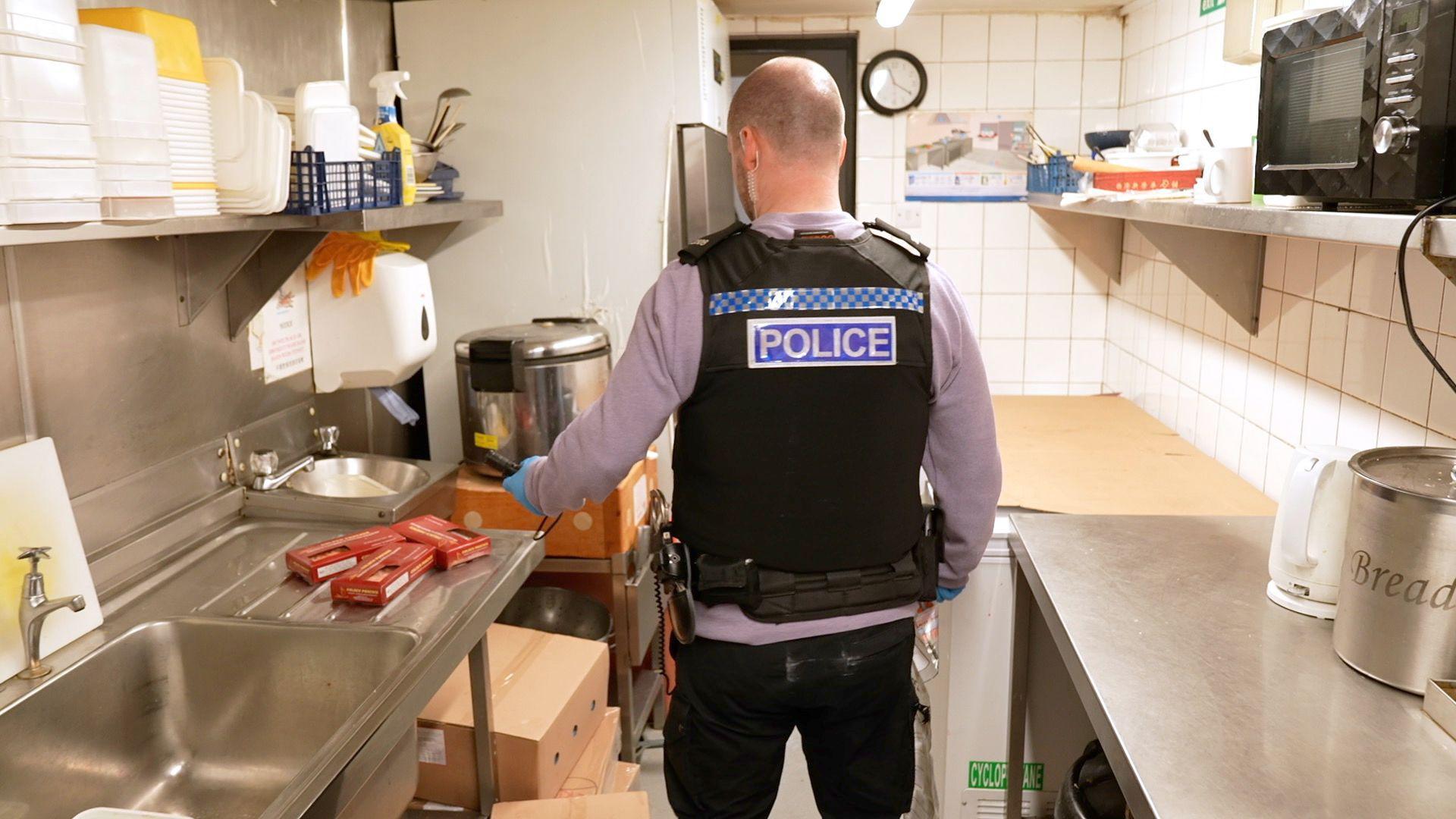 A bald man wearing a grey sweatshirt and a black police stab proof vest walks through a kitchen. 