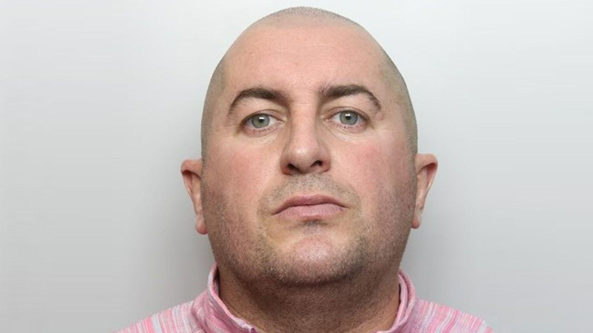 A police mughsot shows a man with a shaved head in a pink top against a grey background.