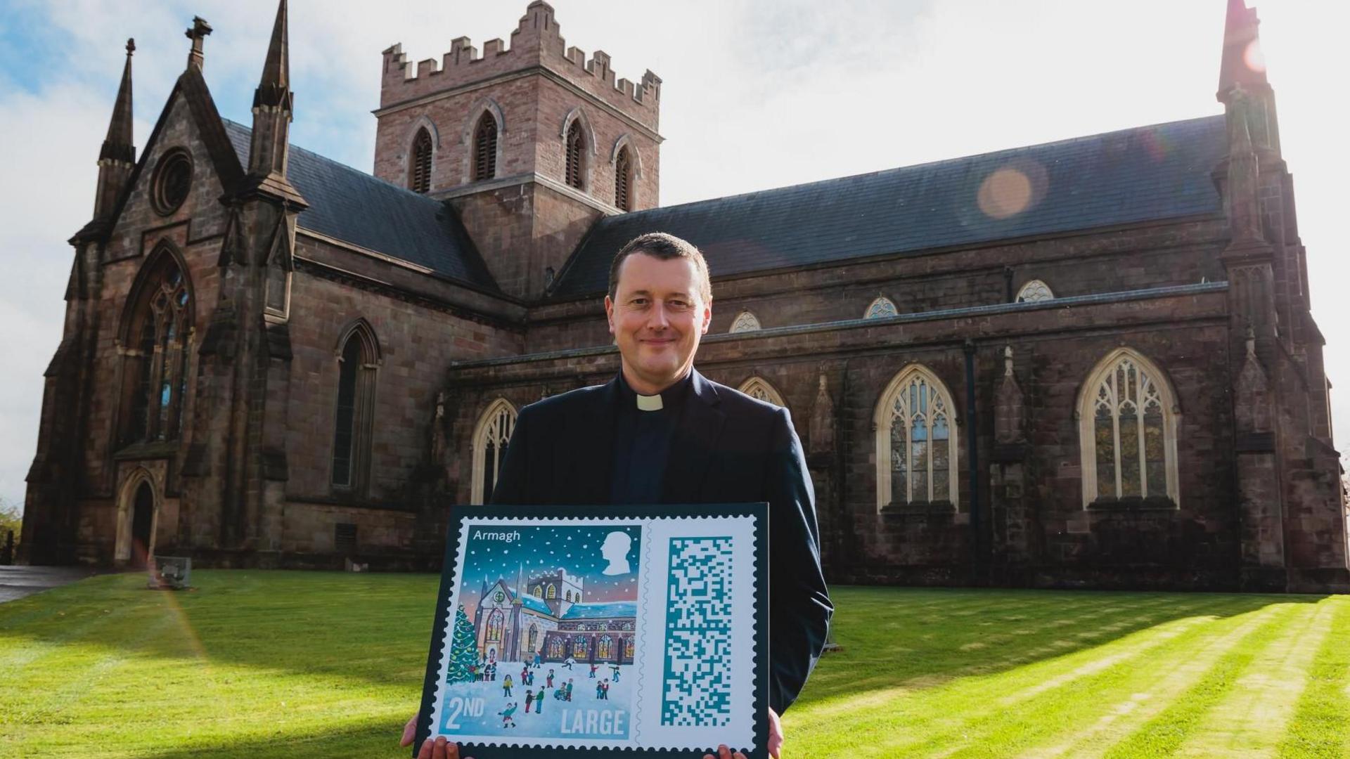 The Dean of Armagh, The Very Reverend Shane Forster, holding a small sign with the image of the stamp design on it - the stamp is an illustration of St Patrick's Cathedral with lots of children outside of it playing in the snow. The cathedral is covered in snow with the lights on and the sky has a dark sunset. The Dean is standing outside the cathedral while holding the stamp. 