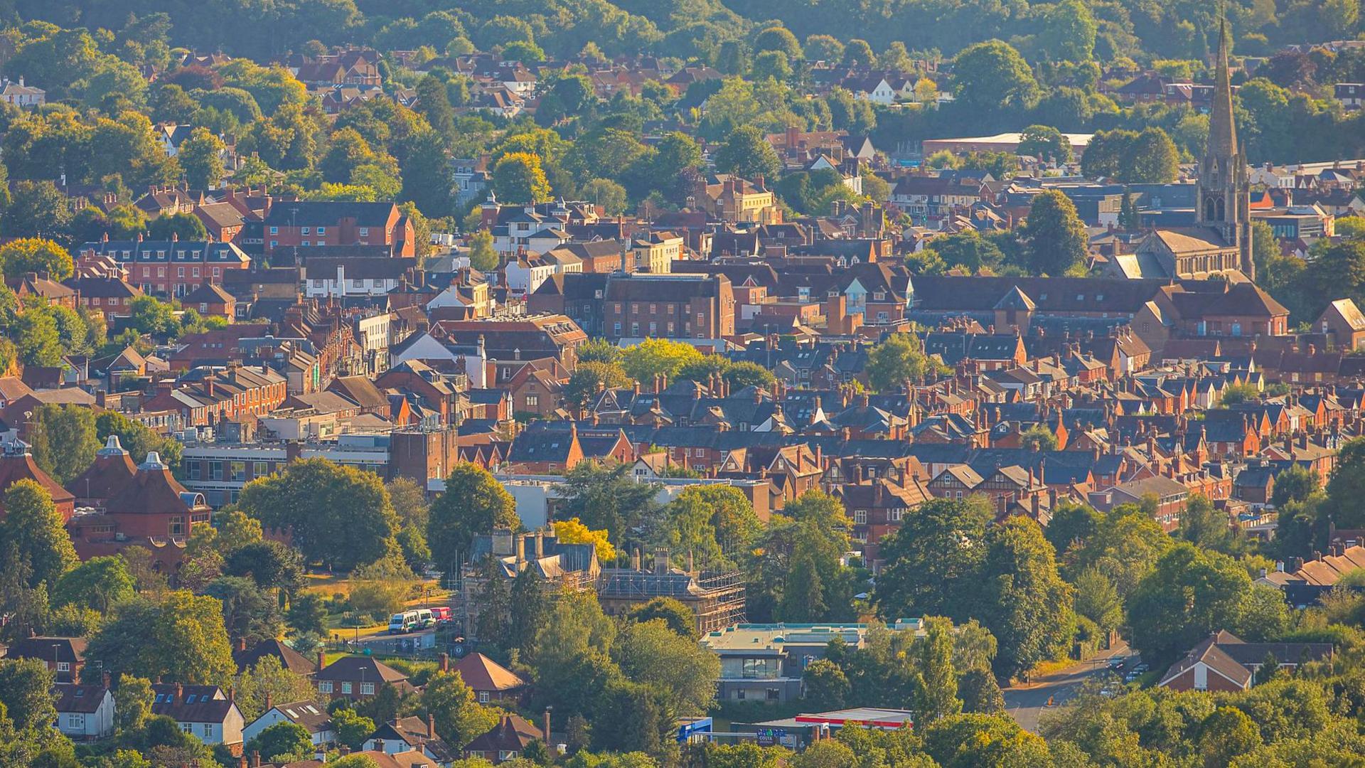 View of Dorking town centre from Surrey Hills. Sun shining on the buildings, in between green trees are lots of houses and an obvious church spire. 