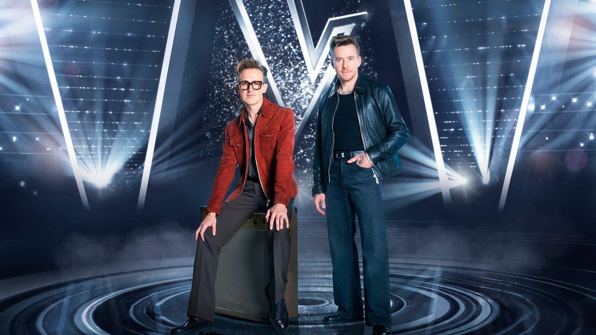 A man in a red jacket with black trousers sitting on an amp alongside a man with a black leather jacket and black jeans standing.
