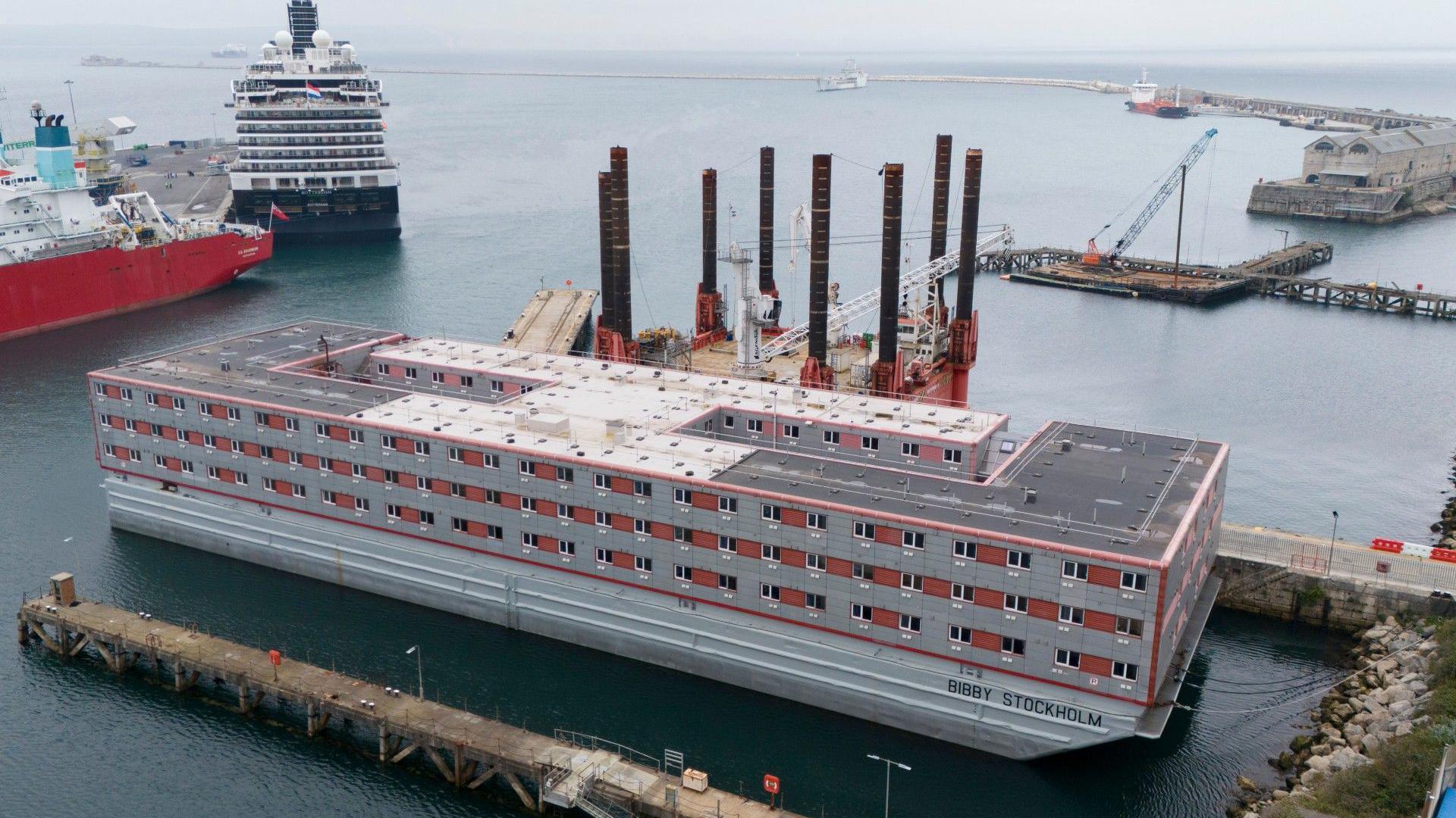 The Bibby Stockholm accommodation barge, with windows on three main floors, is moored alongside a quay at Portland Port. A cruise ship is moored nearby.