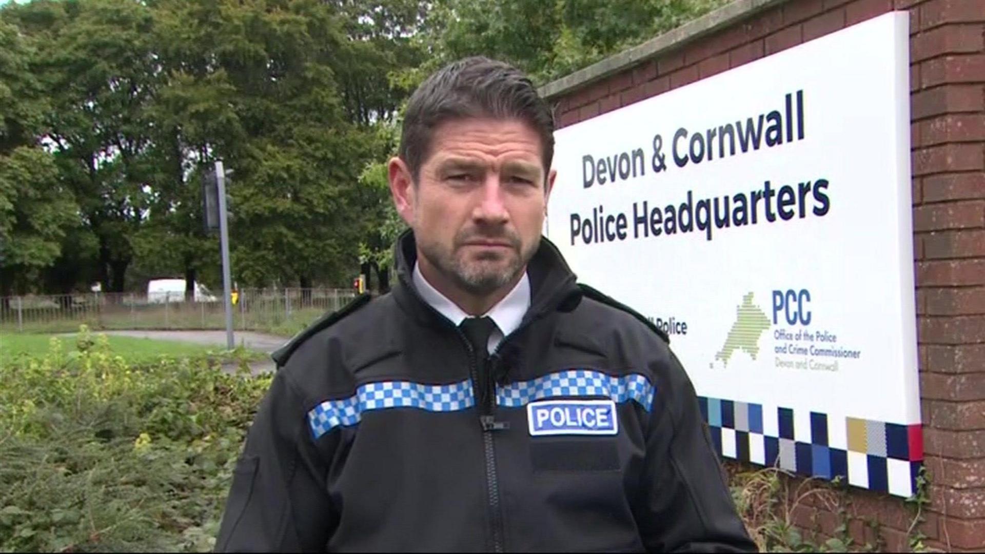 Acting Chief Constable Jim Colwell pictured. He is stood in front of a white sign reading: "Devon and Cornwall Police Headquarters." He has short dark hair that has been swept back. He has a short beard. He is wearing a black police jacket with blue and white check pattering. There is also a blue badge on his chest that reads "police".