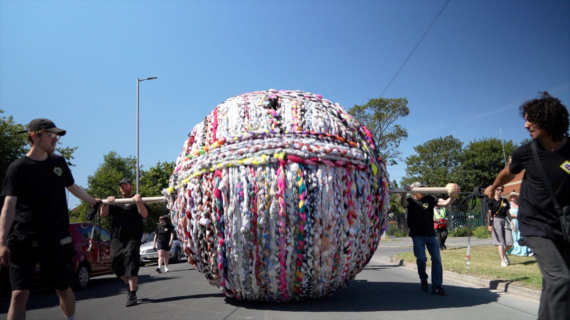 A giant ball of yarn is rolled along a road by people holding a piece of wood which runs through the ball.