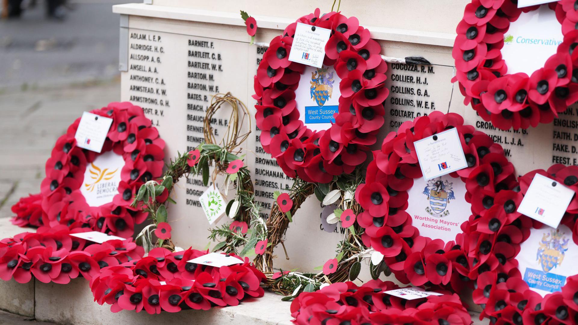 Several wreaths of red poppies lay on a war memorial with black names engraved into it.