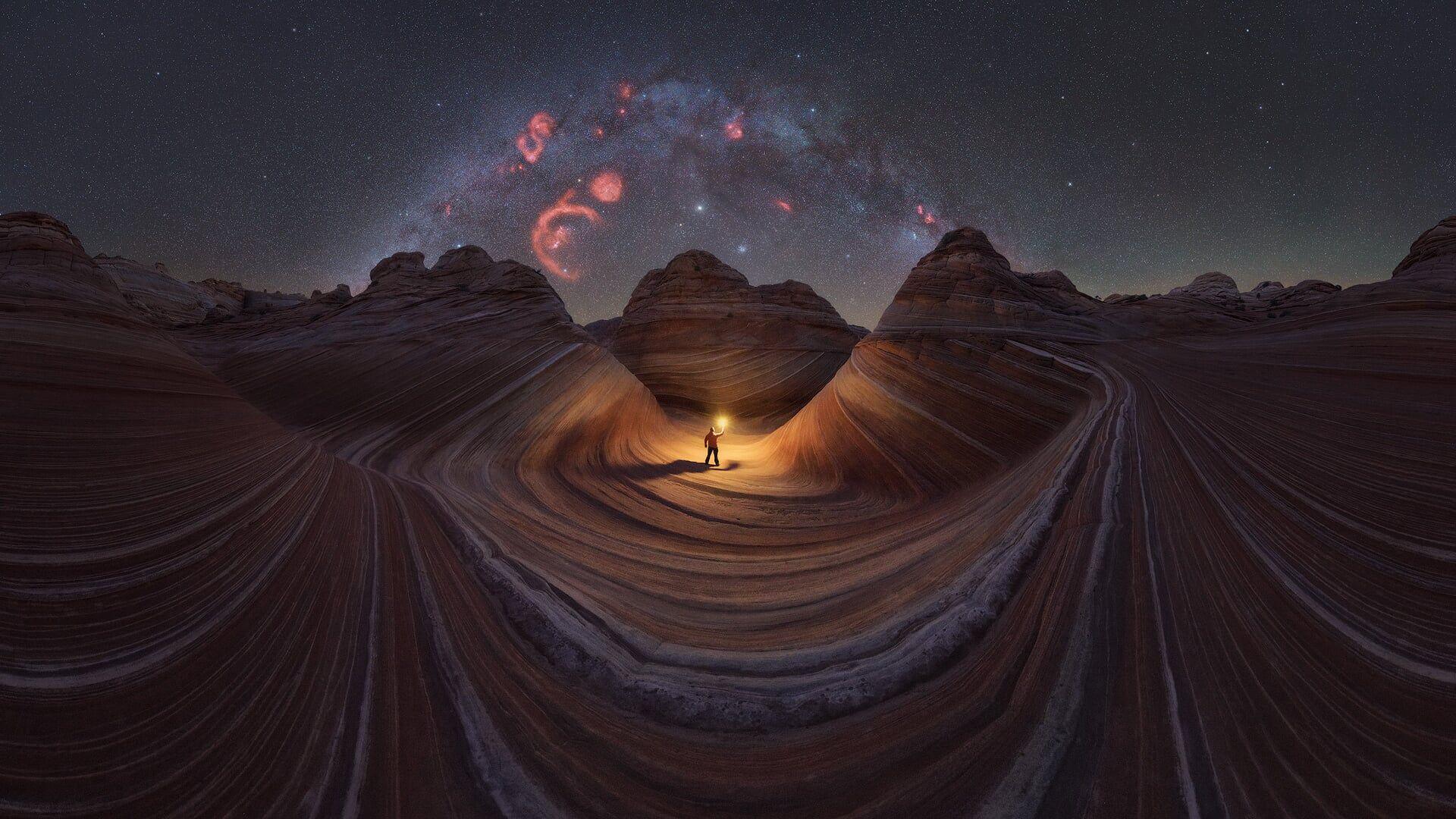 A person holding a light, with sandstone waves around them and with the Milky Way above.