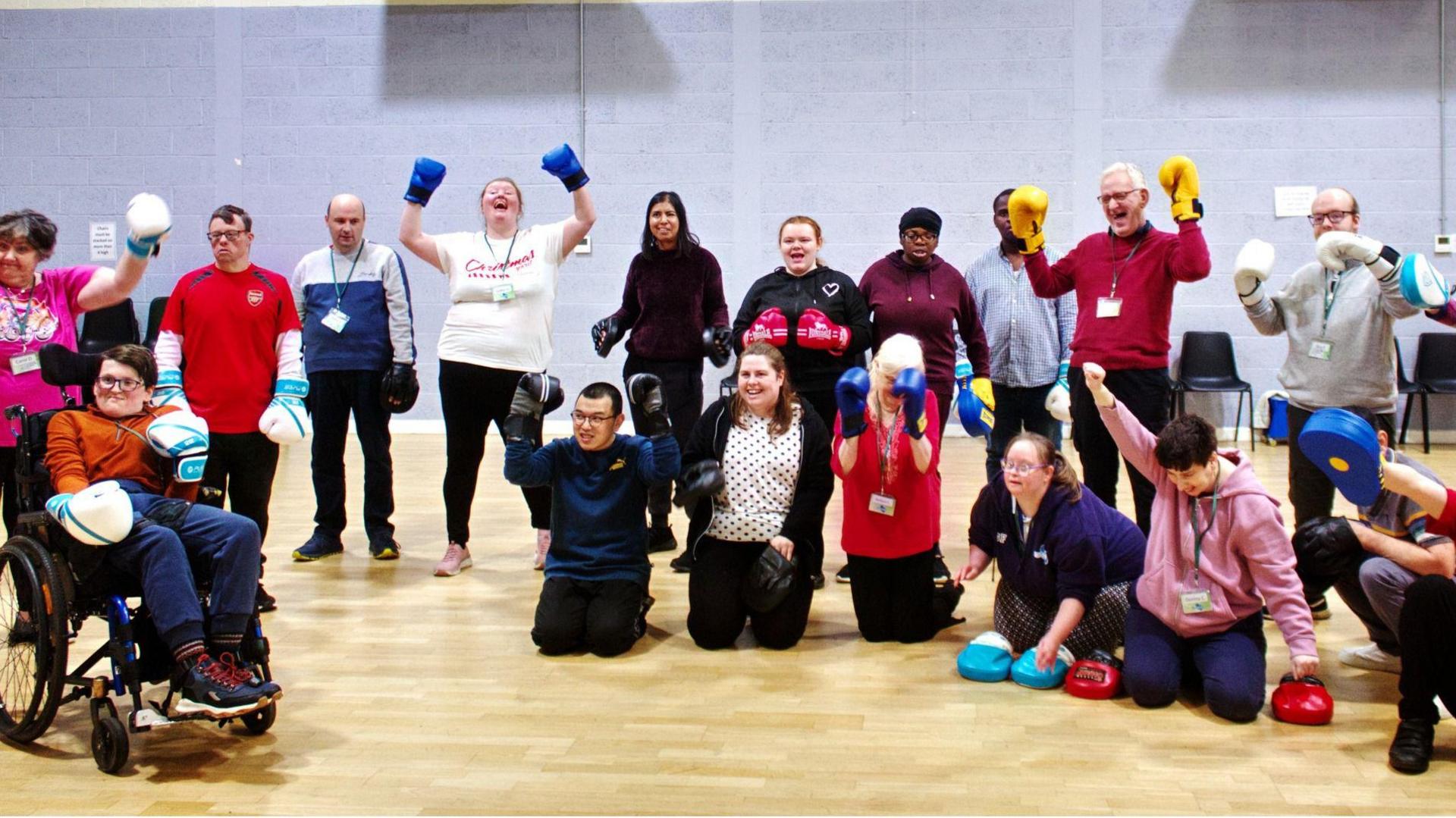 A large group of people from MK Snap. They are all wearing boxing gloves with several holding up their arms and laughing and smiling. One person to the left is in a wheelchair, and six people are kneeling on the floor. They are in a large sports room with a wooden floor and grey walls. 