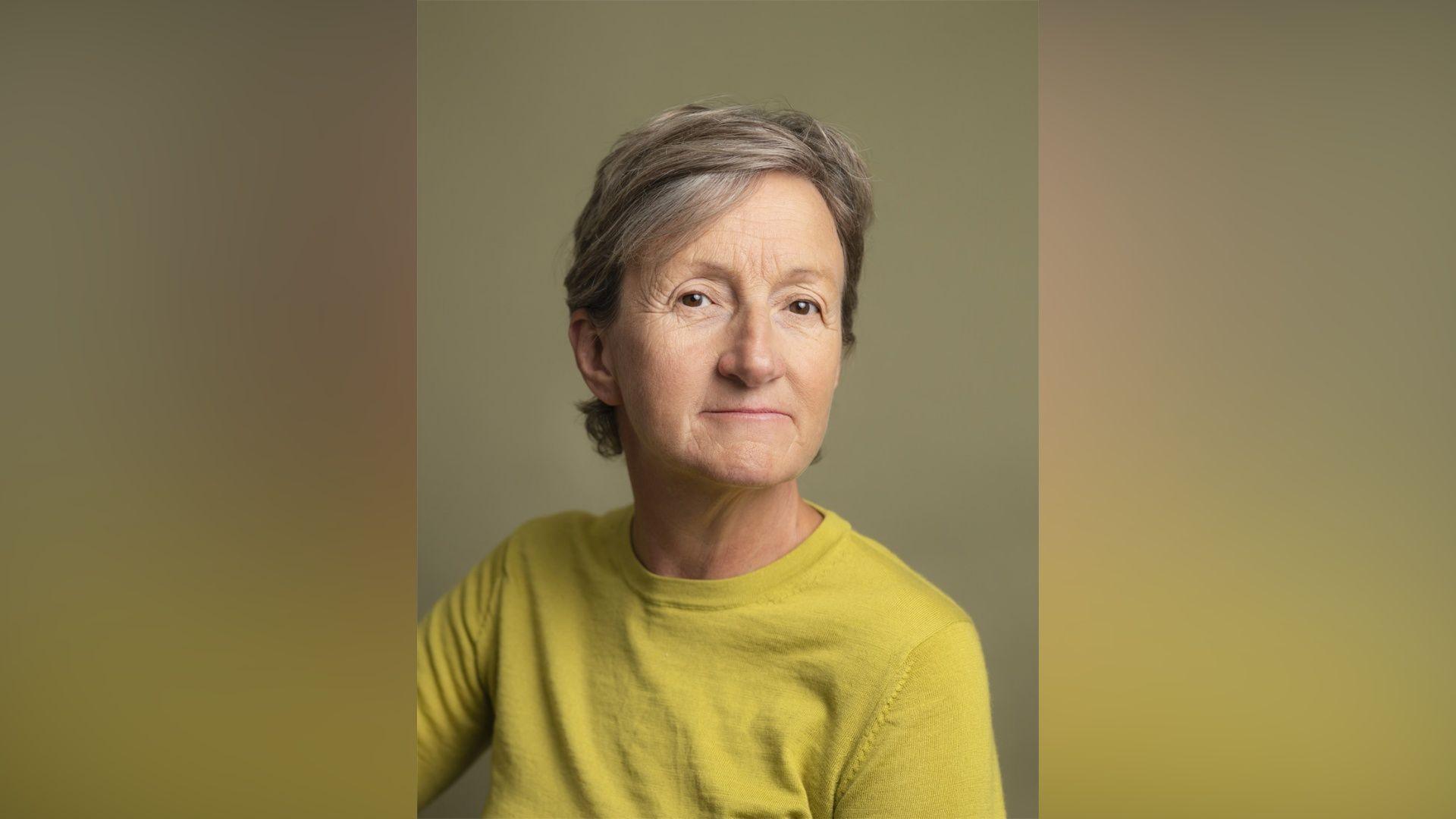 Sarah Hendrickx. She has short grey hair and brown eyes. She is wearing an acid green jumper and looking at the camera with a slight smile.