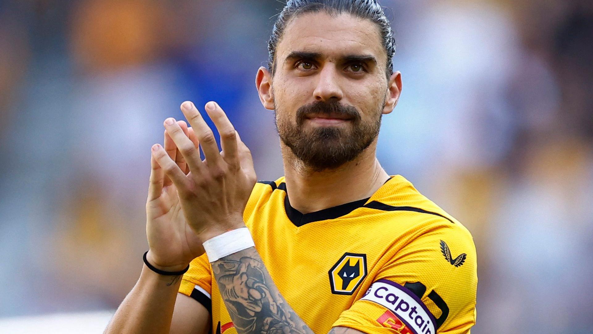 Footballer in a yellow jersey applauding supporters. He is wearing a captain's armband and his right arm is heavily tattooed