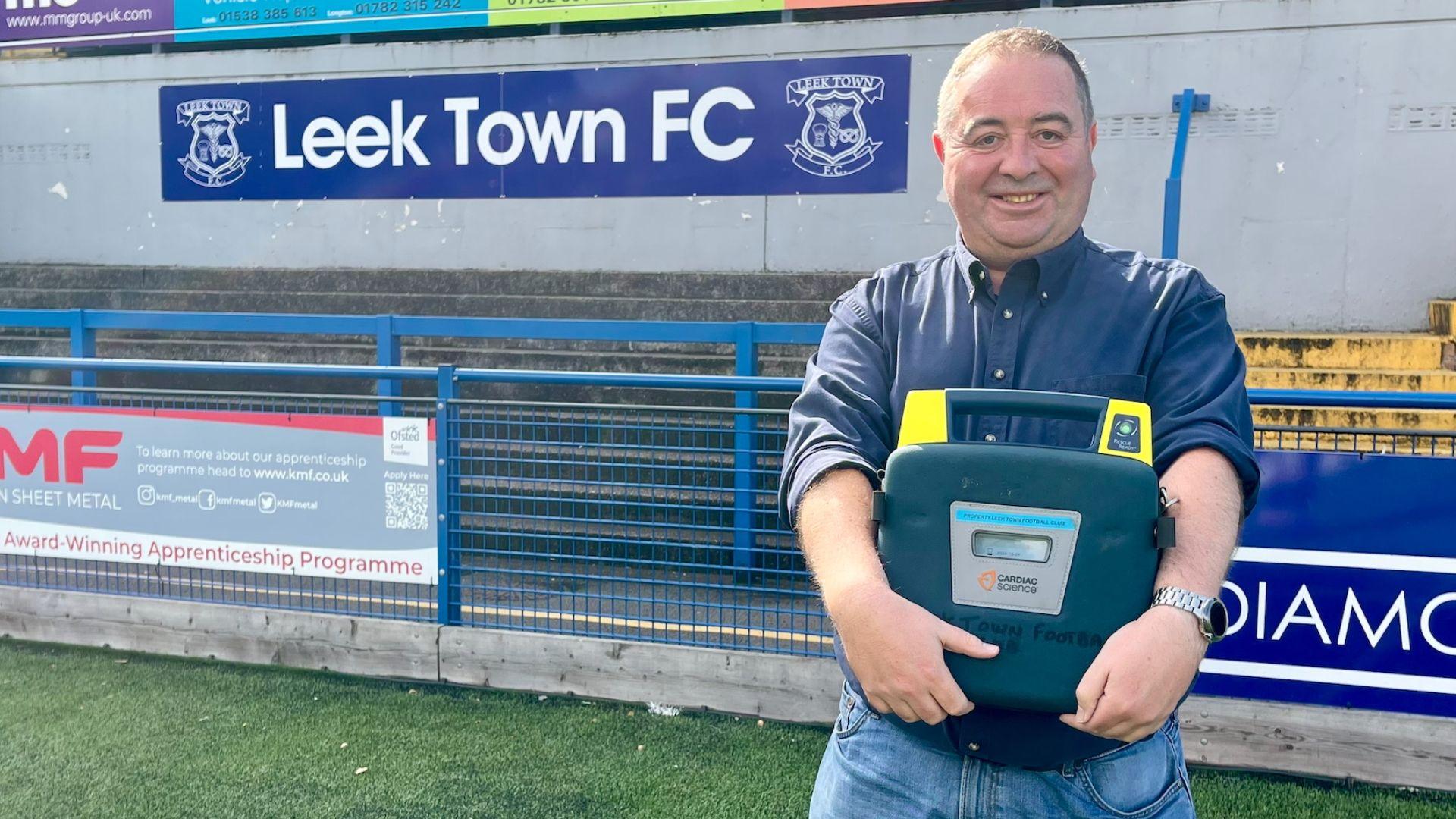 A man with short dark hair is wearing a navy blue shirt and jeans and holding a defibrillator in its case. He is standing on a pitch in front of a football terrace. There is a large blue and white sign that reads, LEEK TOWN FC, with a club crest on either side.