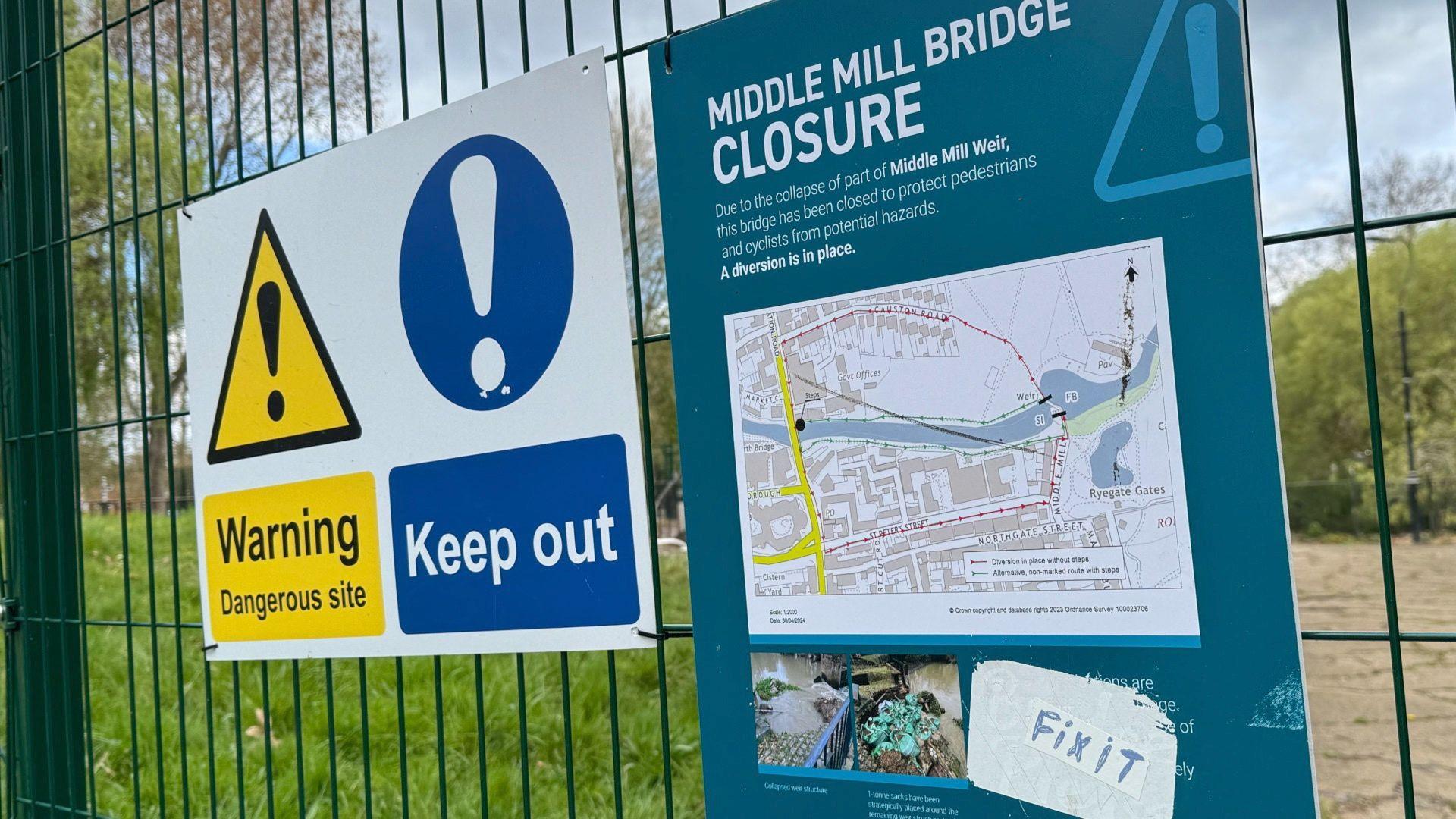 Two signs attached to a green wire fence. On the left is a white rectangular sign with a yellow warning sign above the words "Warning. Dangerous site" and a blue warning sign above the words "Keep out". On the right is a blue sign, about A4 size, with details of a diversion route.
