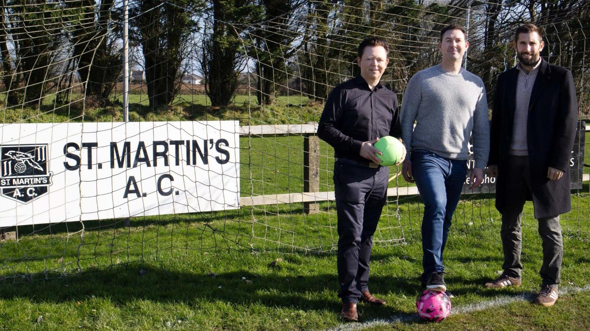 Three men stand in a football goal on a grass pitch - one holding a ball and one standing on one - with a St Martins AC banner behind them.