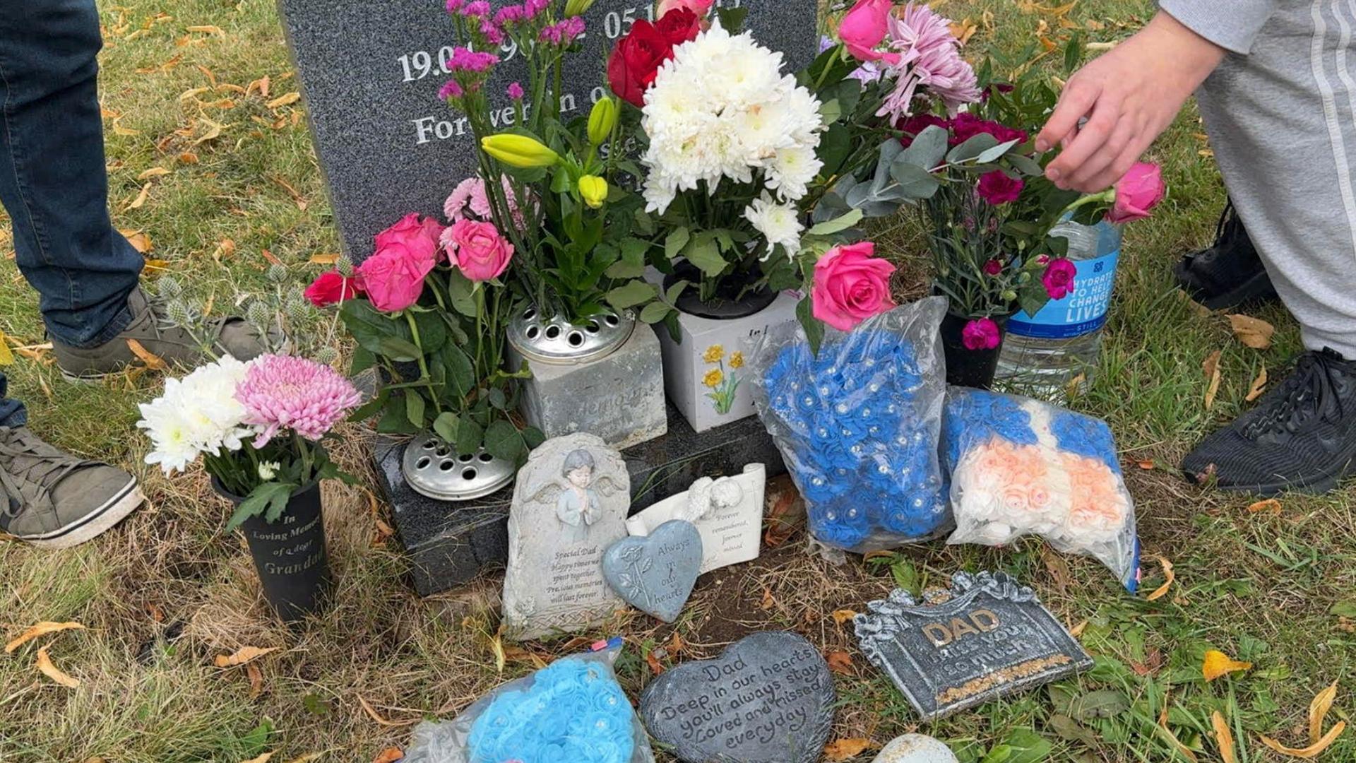 A image of a grey coloured headstone marking a grave. It is surrounded by multiple plaques, bouquets of flowers and plastic bags with flowers.