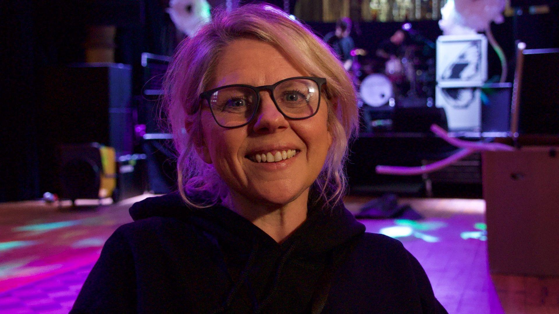 Zoe Alexander has blonde hair, wears a dark jumper and has large dark-rimmed glasses. She sits in front of a stage with purple lighting and drums and amplifiers.