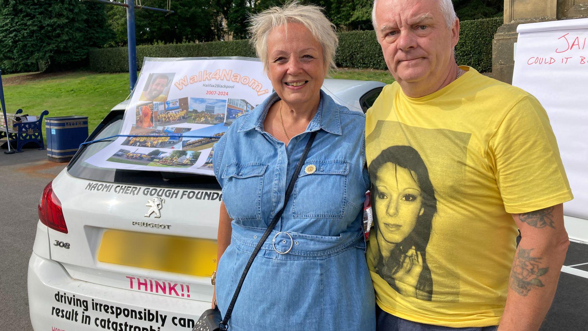 A person wearing a yellow t-shirt with a picture printed on it standing in front of a car with writing on