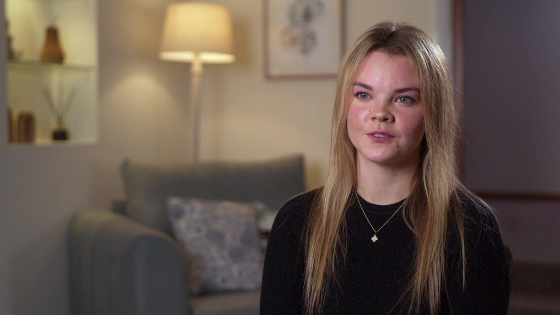 Eloise Cosser - an 18-year-old with long blond hair wearing a dark top and a chain in a room with a chair and light in the background