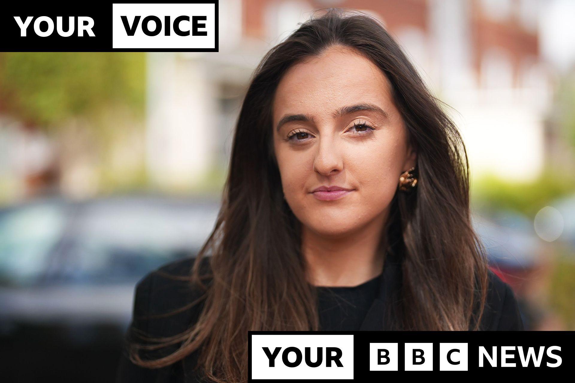 A woman with flowing long brown hair, wearing a black jacket and black top looks pensively into the camera. Behind her are several cars out of focus. Overlaid on top of the photo is a black and white graphic reading 'your voice, your BBC News' in caps.