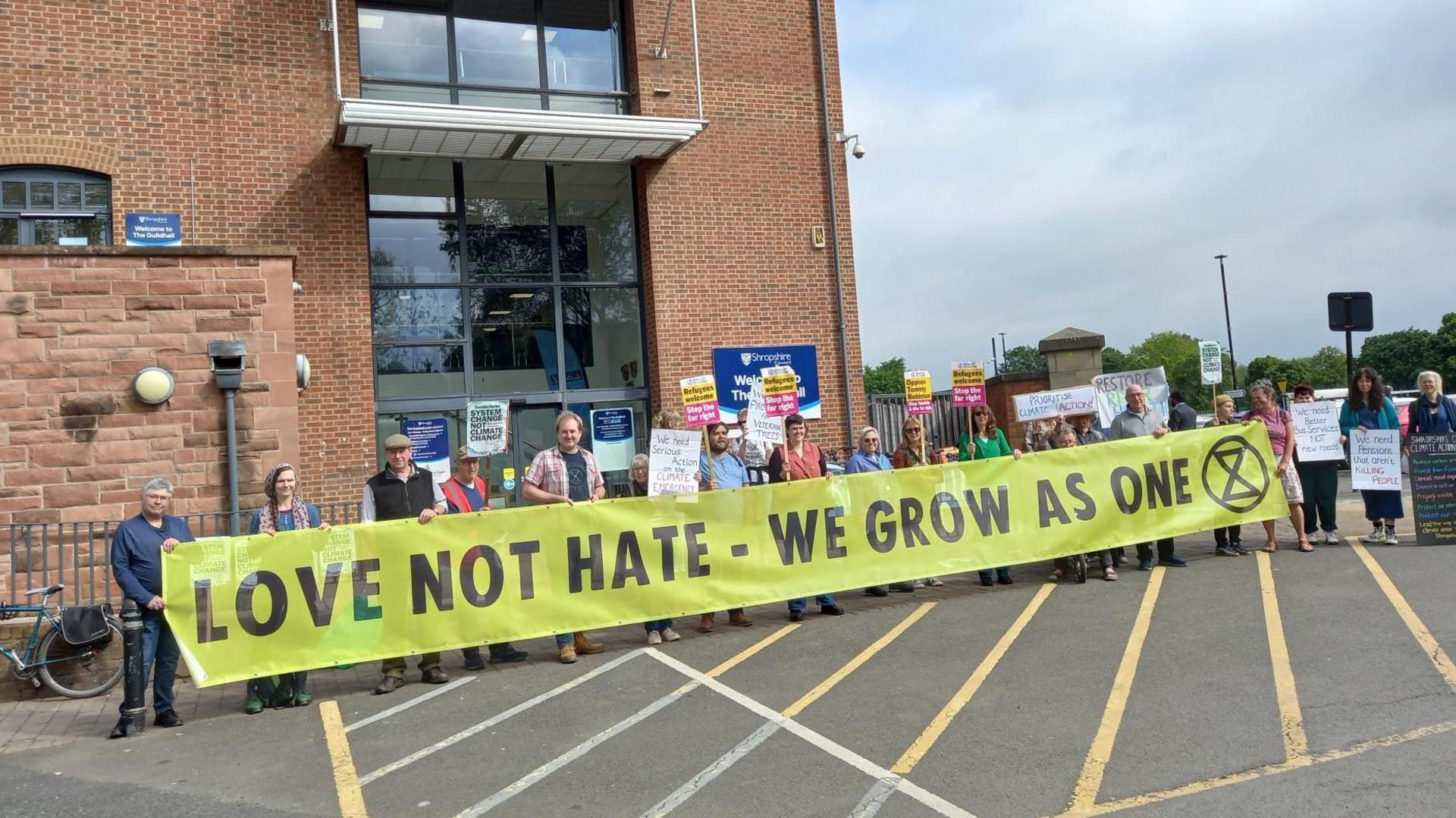 A group of about 20 people holding a yellow banner with black words on it, reading 'Love Not Hate - We Grow As One' and the Extinction Rebellion hourglass logo. They're stood outside a large red brick building with big windows. The sky is cloudy.
