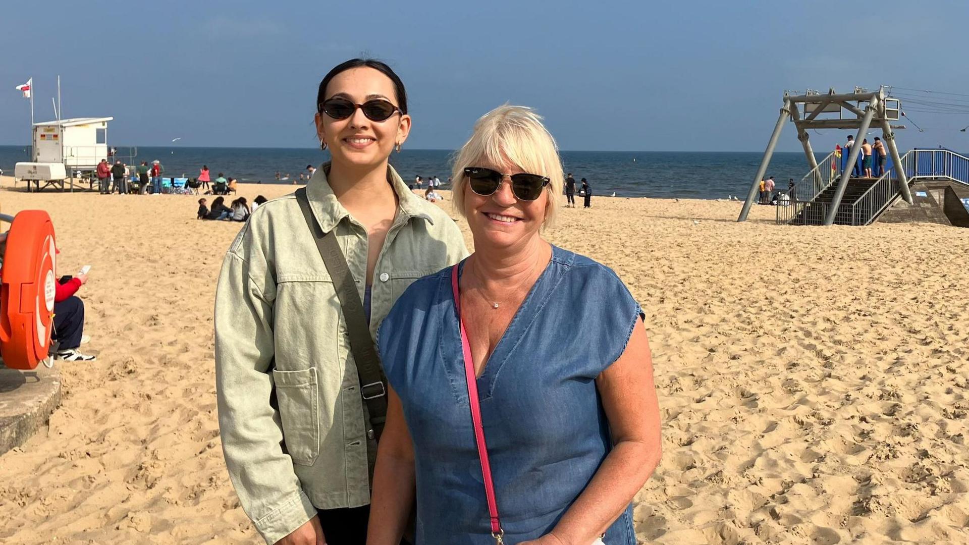 Lauren on the left and Caroline on the right standing on Bournemouth beach. Lauren has dark hair and is wearing a light coloured jacket and sunglasses. Caroline has blonde hair and is wearing a blue top and sunglasses.