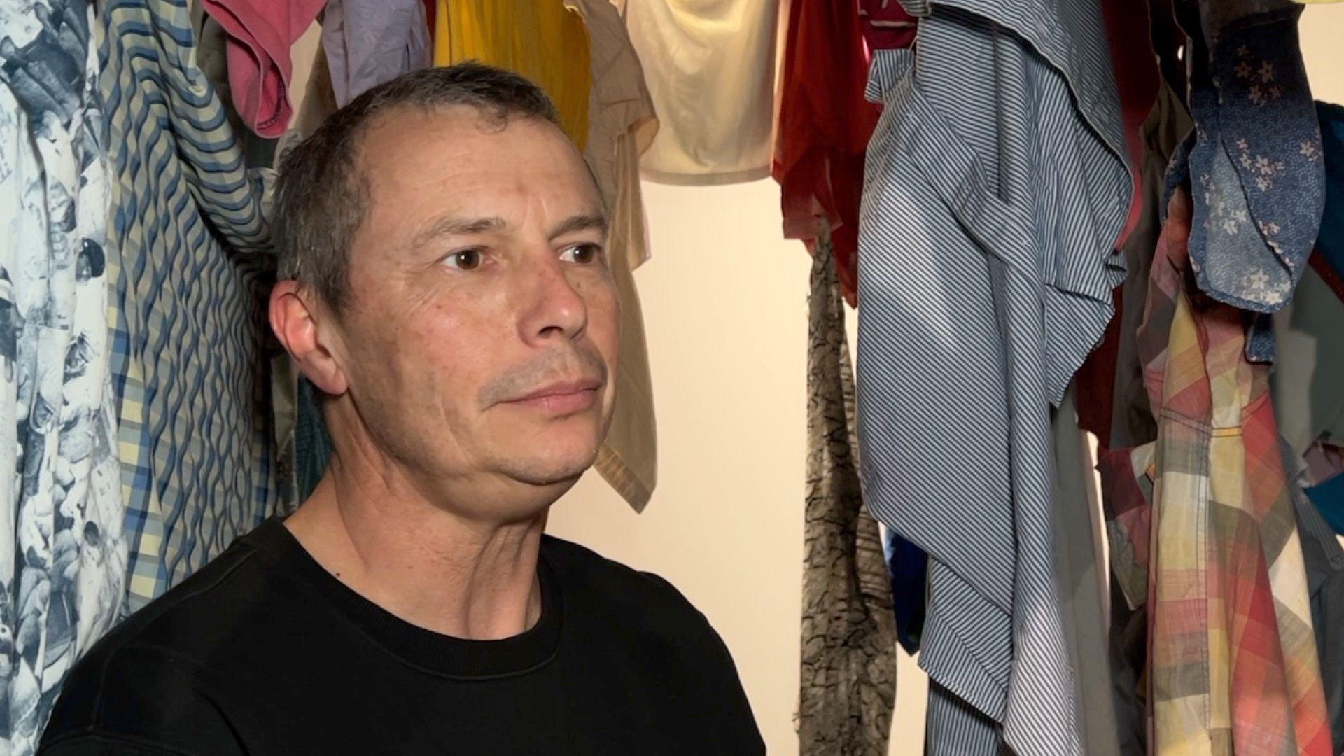 Bruce is standing by shirts that are hanging around him. He's looking to right of the photo and is wearing a black jumper. He has dark eyes and dark hair which is starting to turn grey.