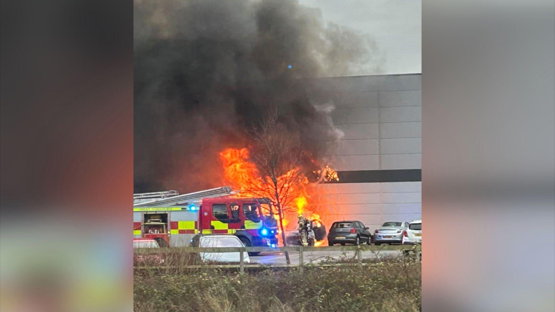 A fire engine with crew tackling the blaze. Black smoke is drifting over a retail park store. 