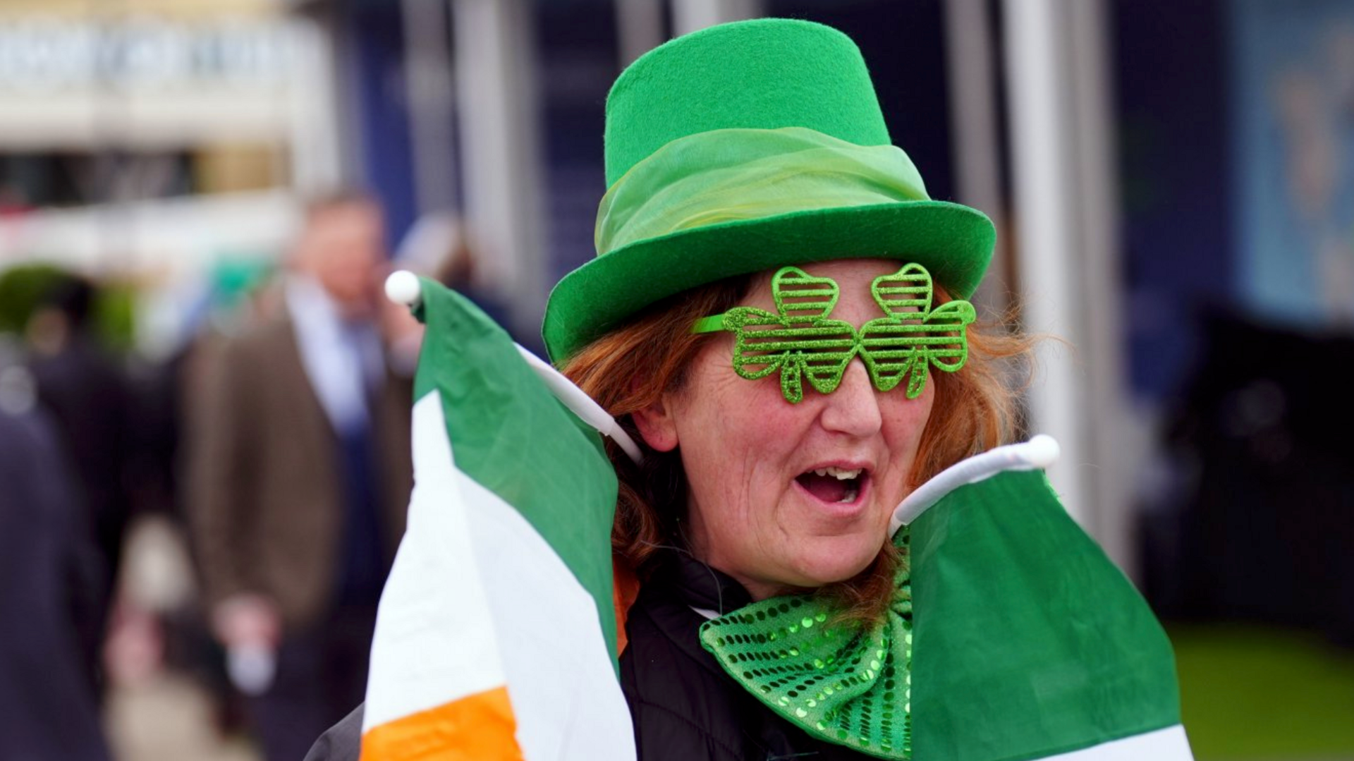 A woman smiling and wearing a green hat and glittery green four-leafed clover glasses. She has red hair and is carrying two Irish flags around her neck.