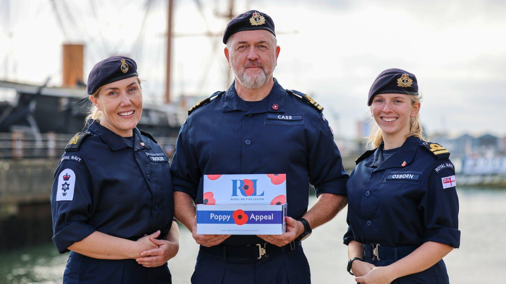 Three members of the Royal Navy stand together to present a box of poppies. A man stands in the centre, holding a white and blue poppy appeal box, two blonde women stand either side of him, with their hands clasped together and smiling. They are all wearing Royal Navy combats - a dark blue shirt and trousers with a beret.