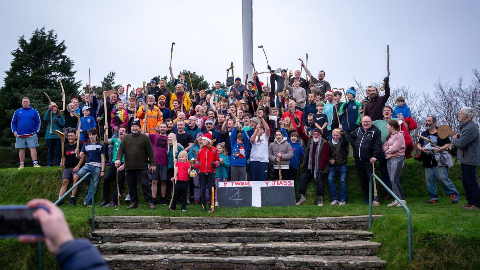 Player from both team gathered on top of Tynwald Hill holding sticks aloft. There scoreboard at the front of the crowd.